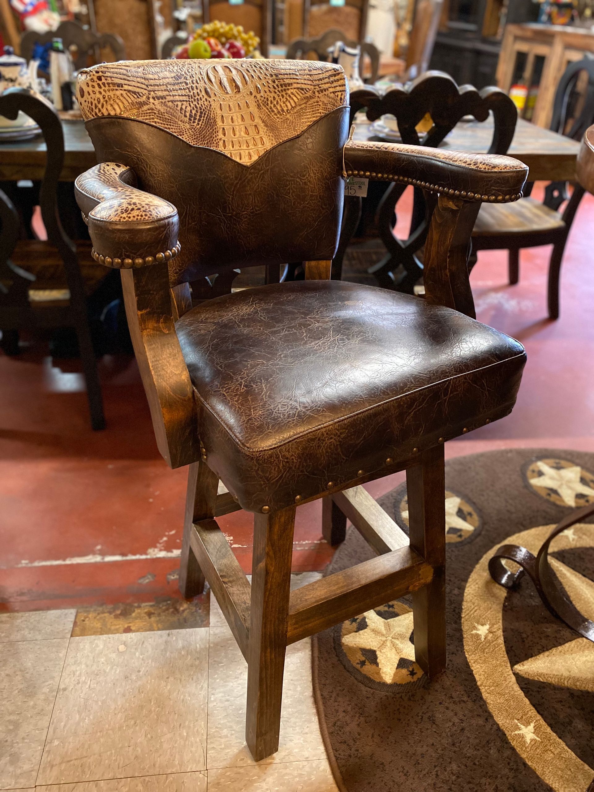 Western-style bar stool with dark brown leather seat, back, and armrests. It has a wood frame and stands in a room with a rug.