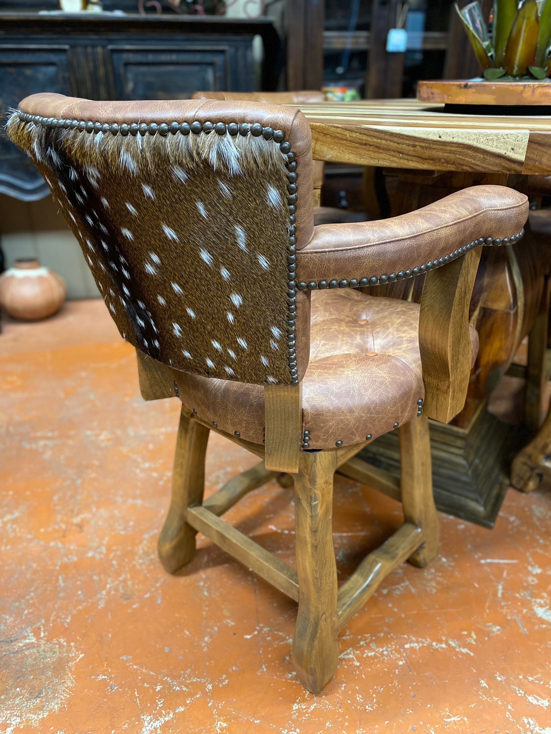 A rustic barstool with a cowhide back and leather seat, brown wooden frame. It's set in a room with other wooden furniture.