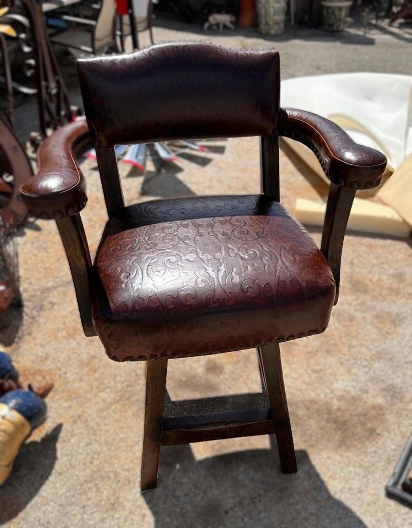 Brown leather-upholstered armchair with curved arms on a wooden frame. The chair is outdoors on a paved surface.