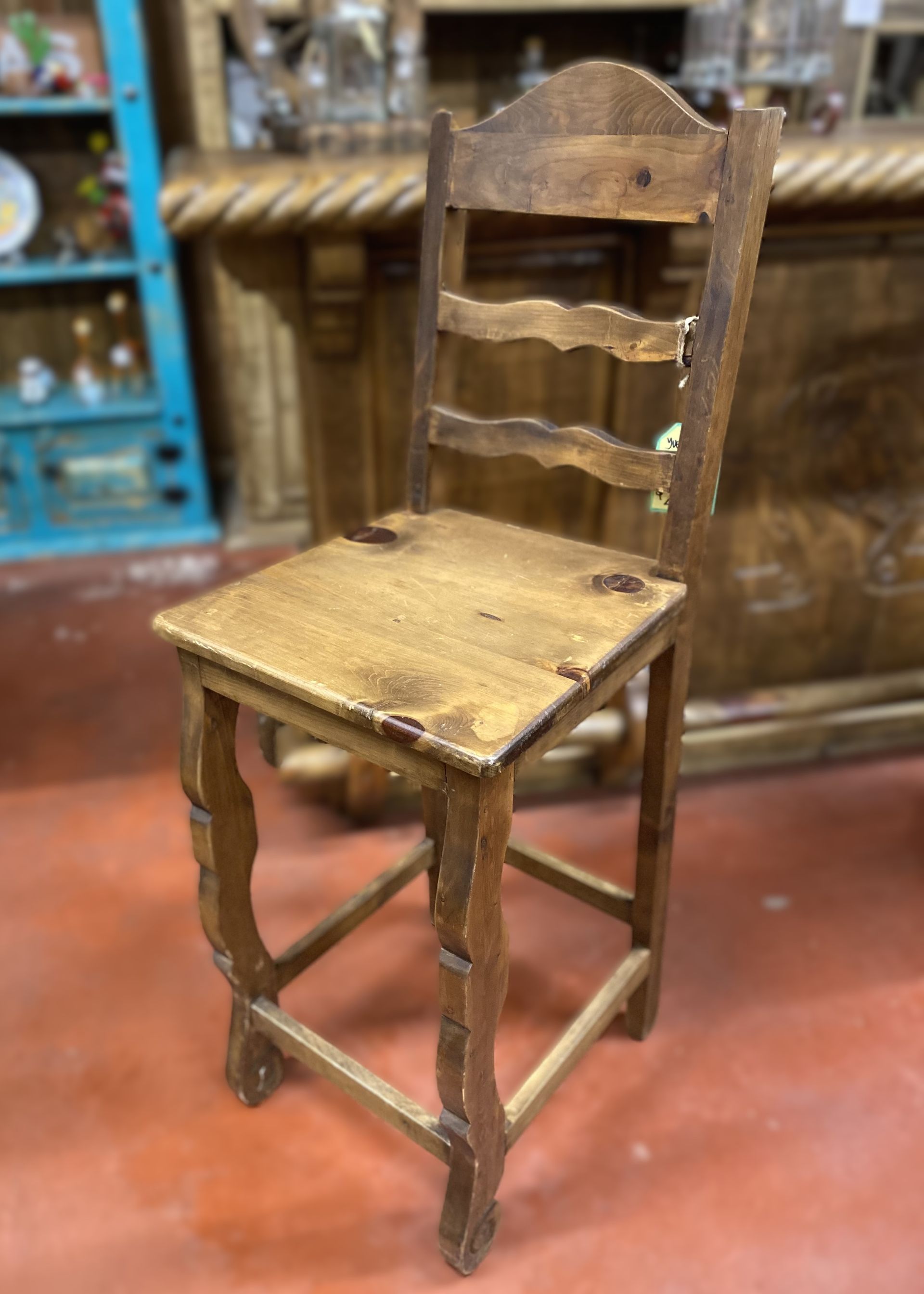 Wooden bar stool with carved legs, a square seat, and a back with three horizontal slats. The setting is a rustic interior.