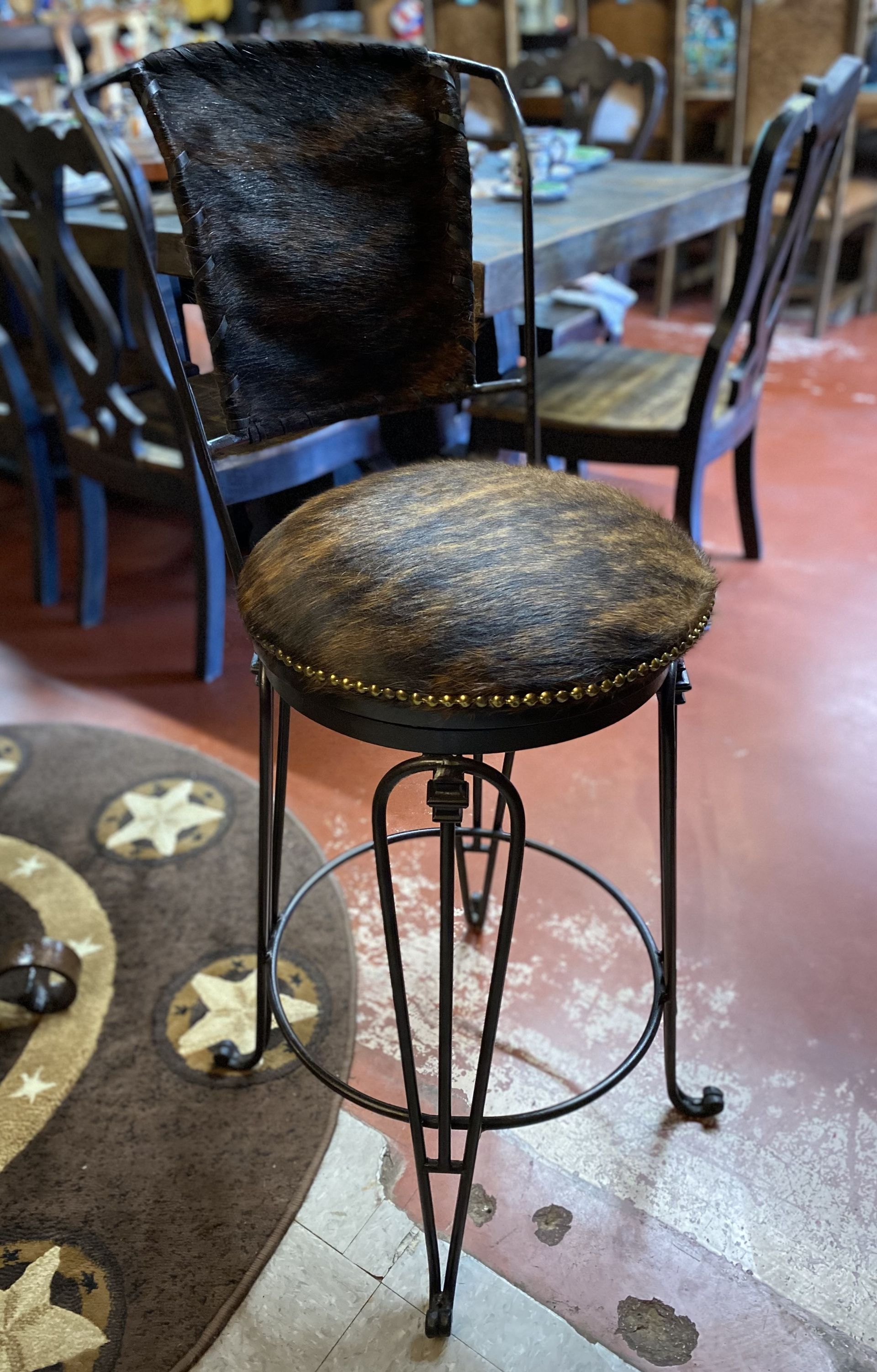 Rustic metal bar stool with a cowhide seat and back. It sits on a patterned rug, with a dining table and chairs in the background.