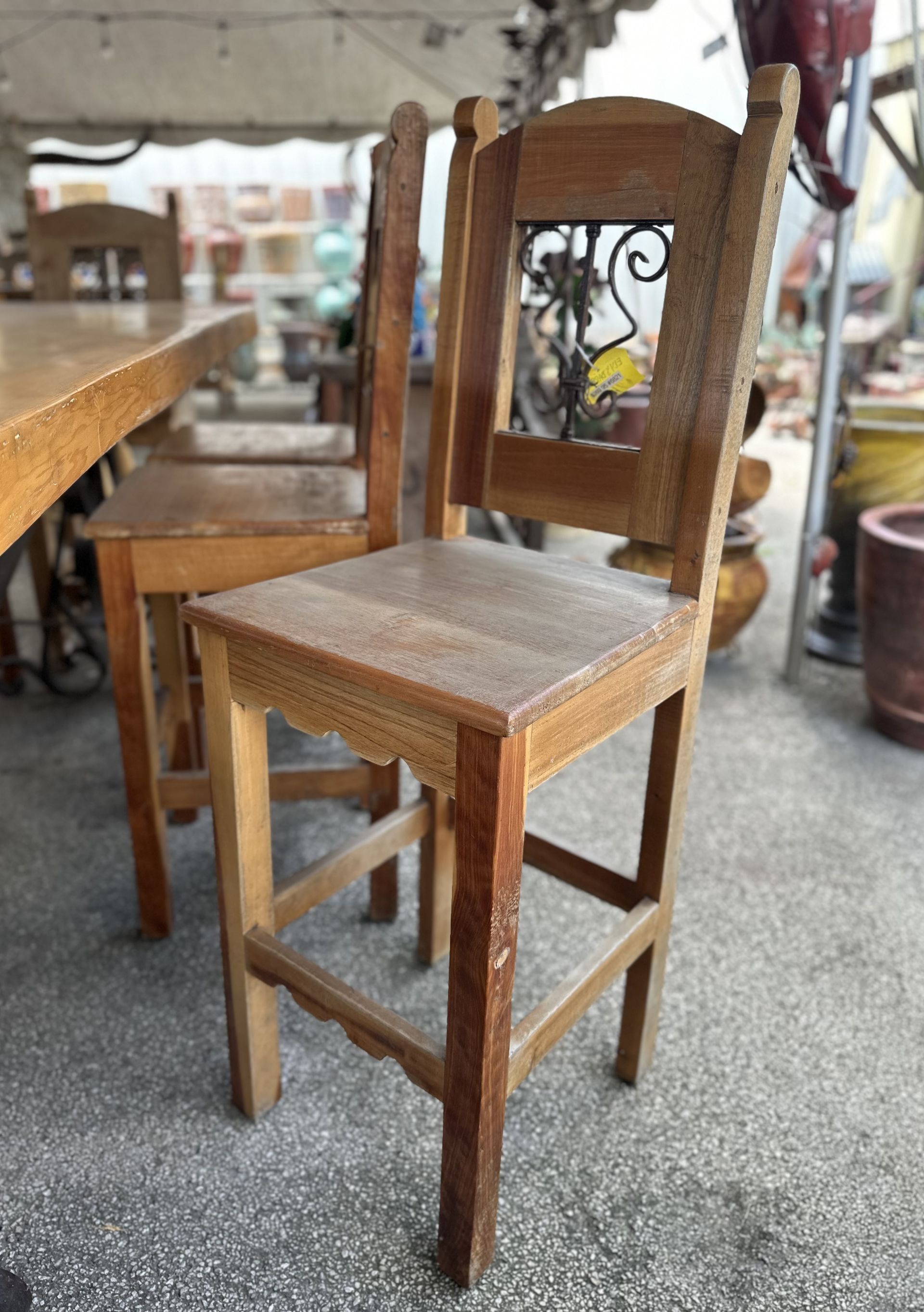 Wooden bar chairs in a light brown color, with a carved back and square seat, stand in an outdoor setting.