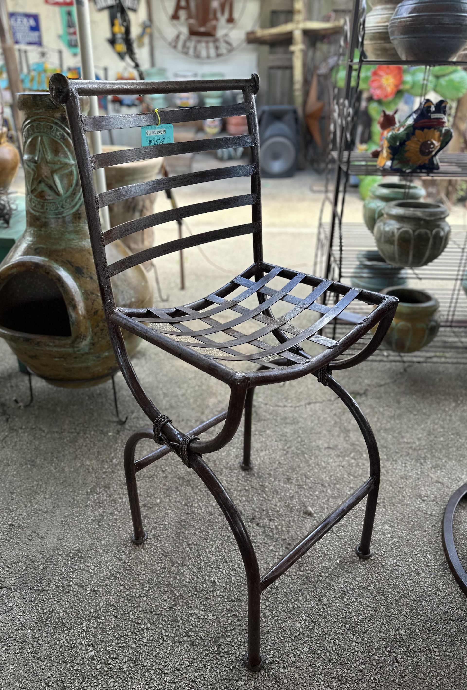 Wrought iron chair with a woven seat and back, brown metal, set on a textured gray surface, outdoors in a shop.