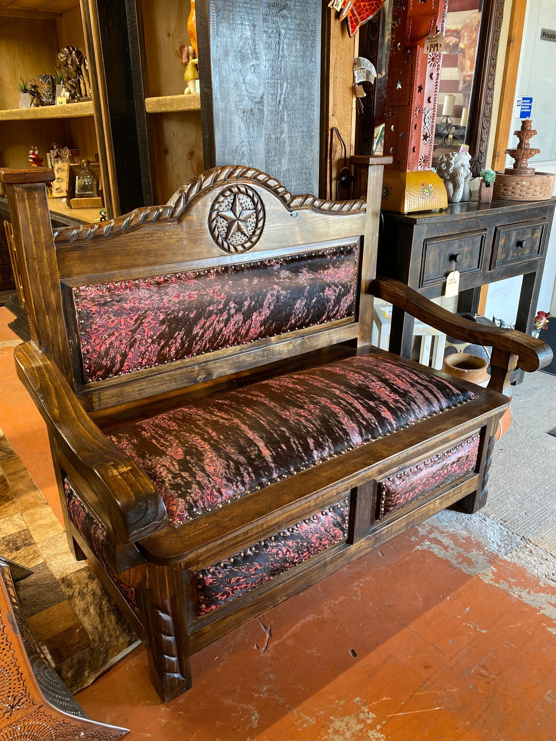 Rustic wooden bench with red and black patterned upholstery. Features a star carving and ornate wooden arms, set in a cluttered shop.