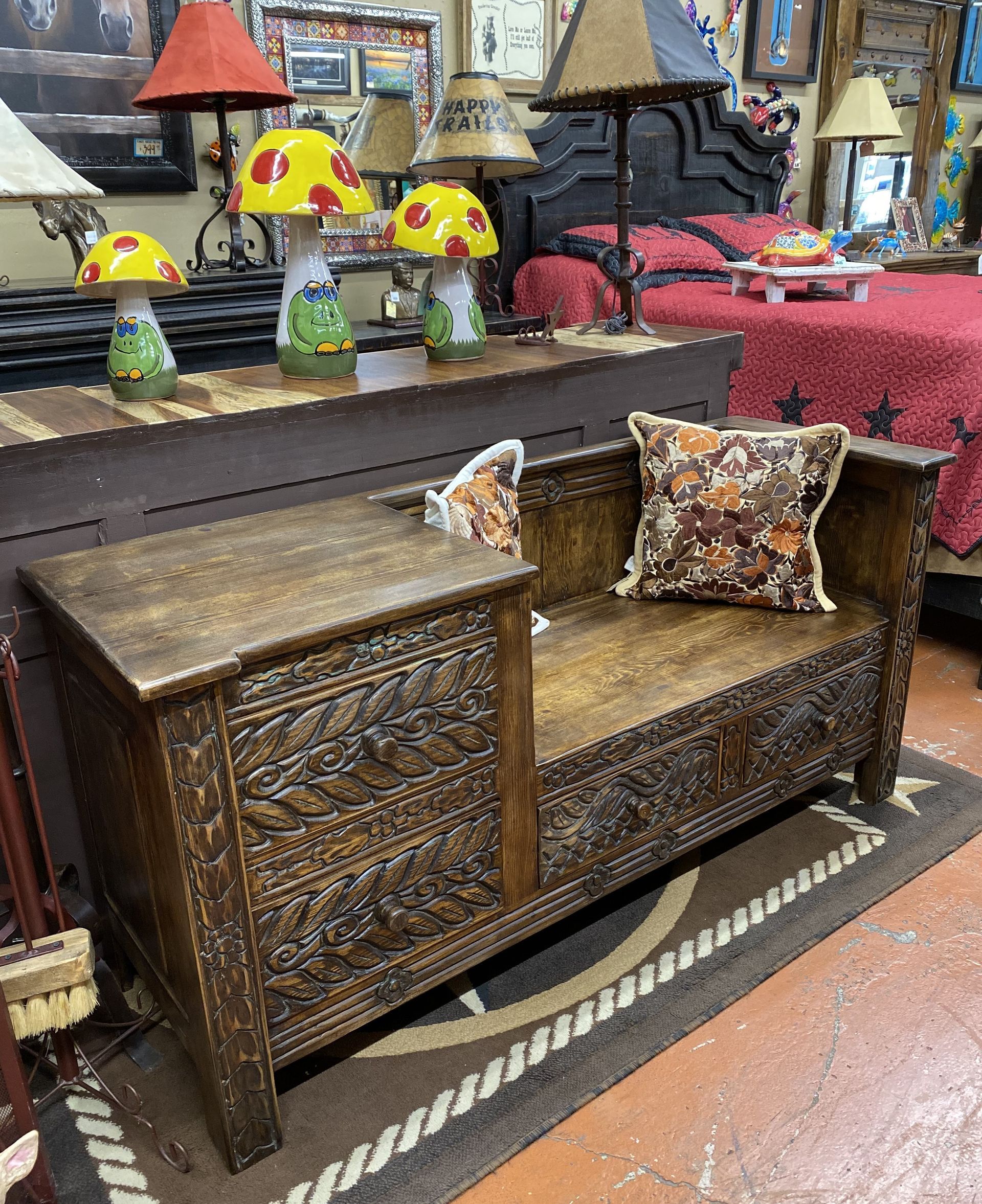 A carved wooden bench with an armrest, a pillow, and detailed floral patterns in a shop setting with lamps and decorative items.