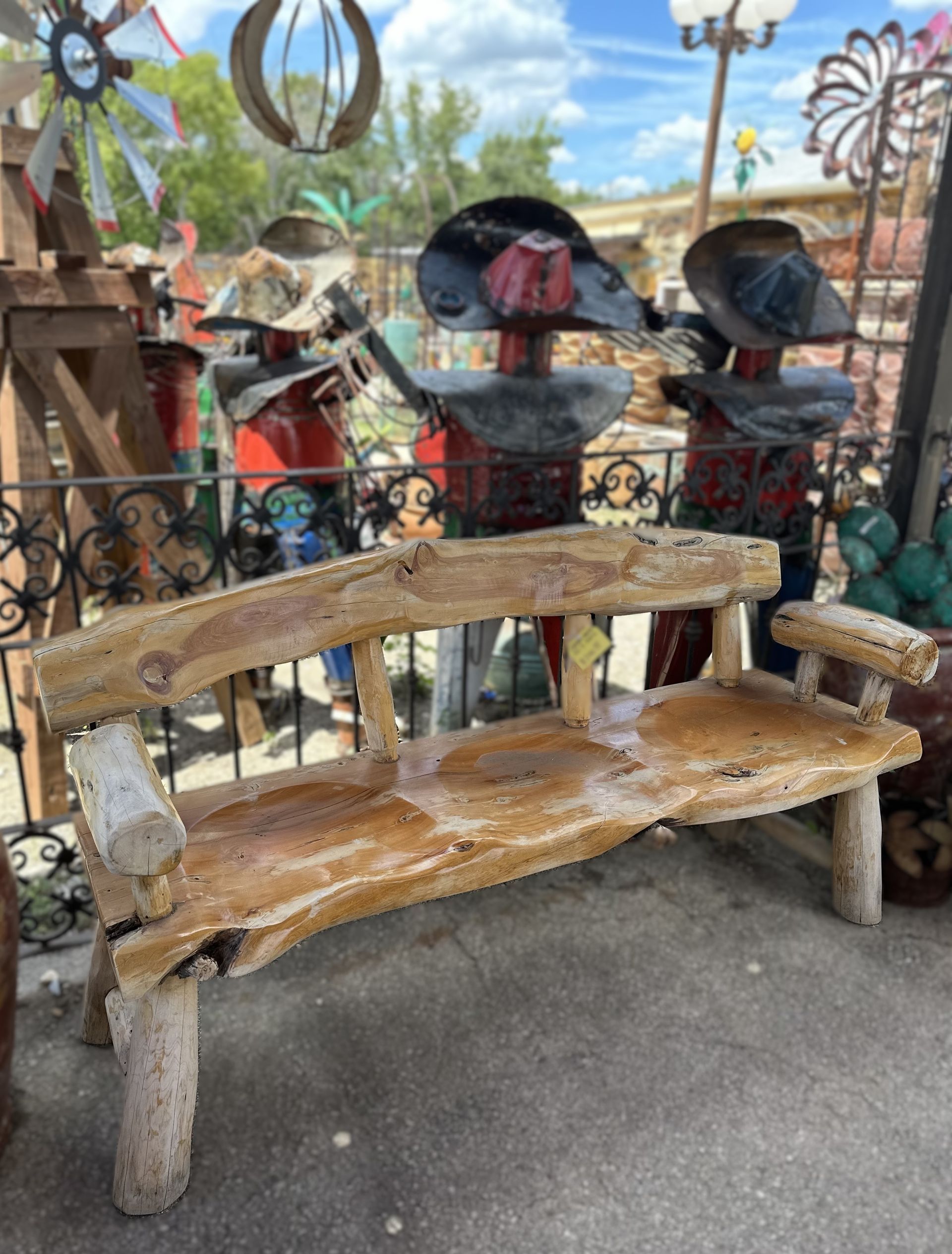 Wooden bench in front of metal art figures wearing hats. The setting appears to be outdoors, possibly a shop or yard.
