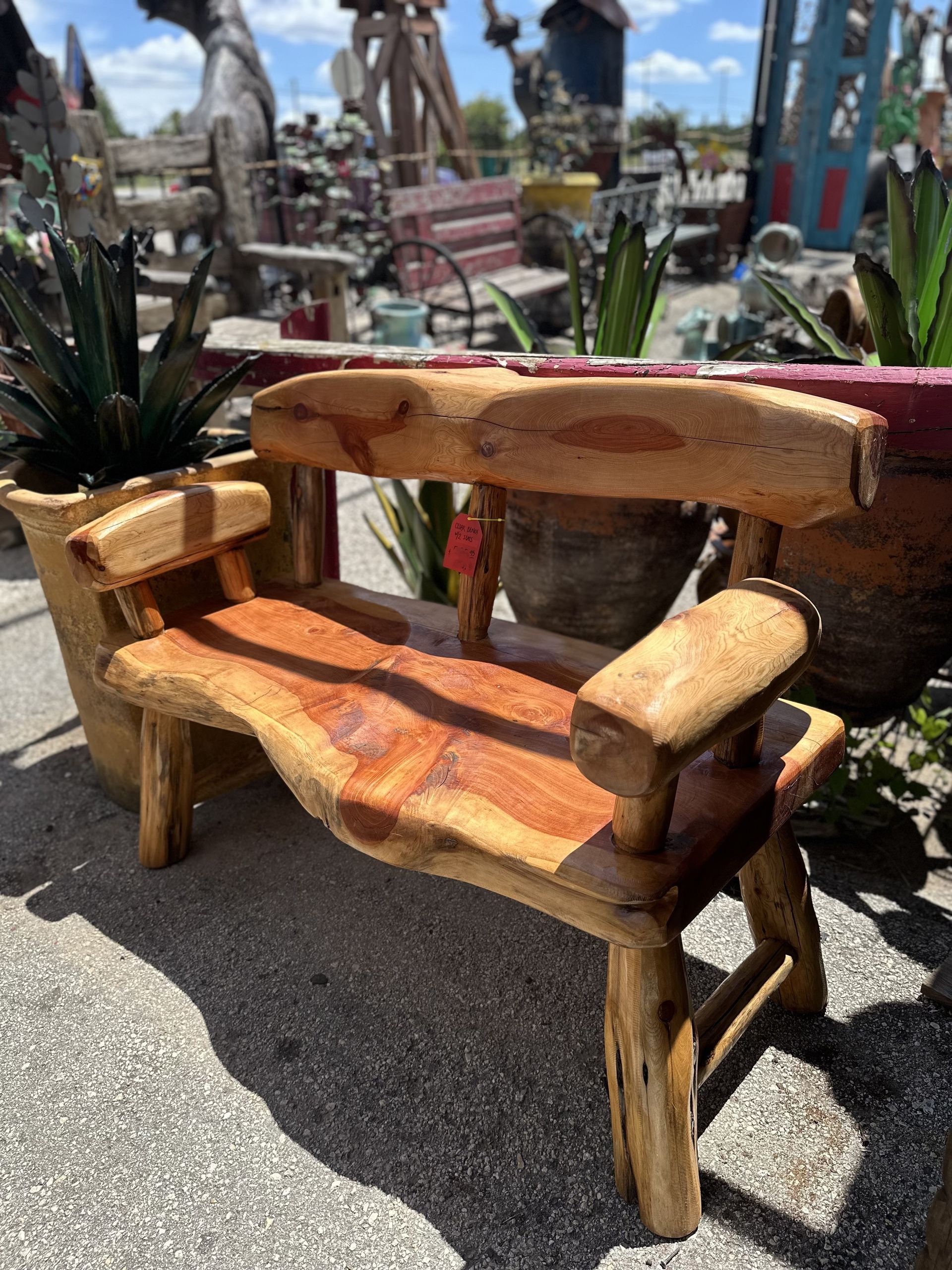 Wooden bench with natural edges, in an outdoor setting with other sculptures and plants, under a sunny sky.