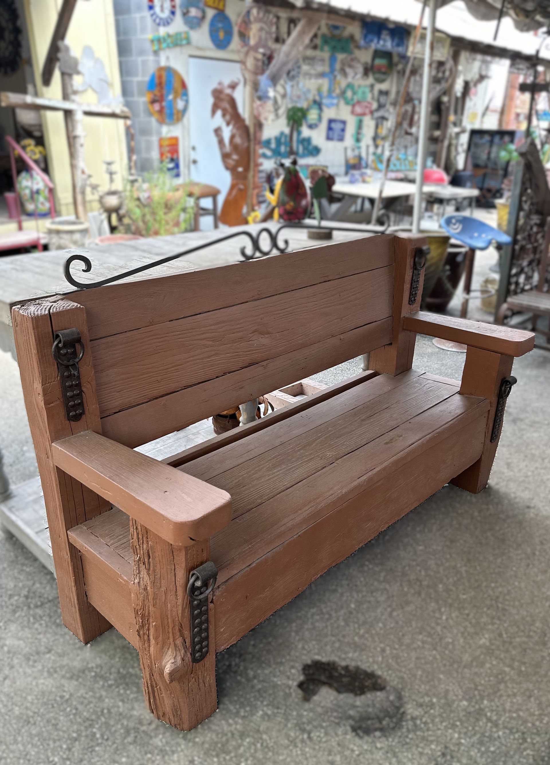 Wooden brown bench with metal accents, outdoors. Features a backrest, armrests, and decorative ironwork above.