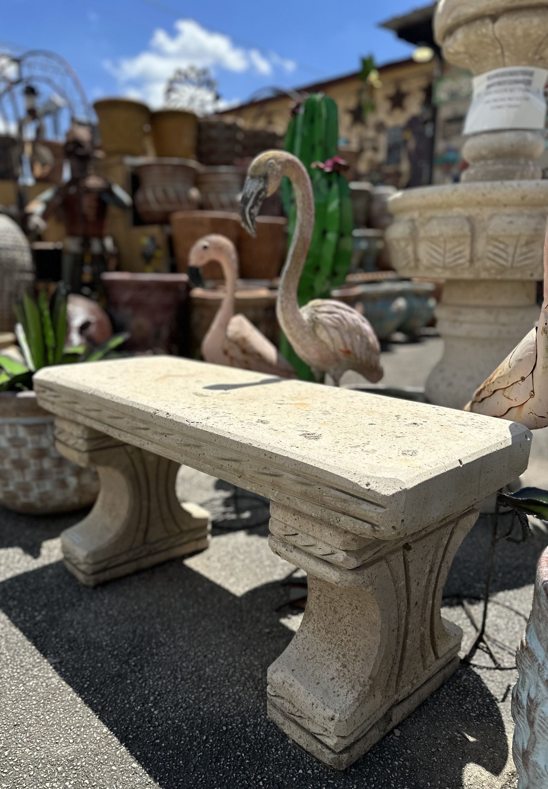 A stone bench with flared legs sits in an outdoor garden center, with flamingo statues and various planters in the background.