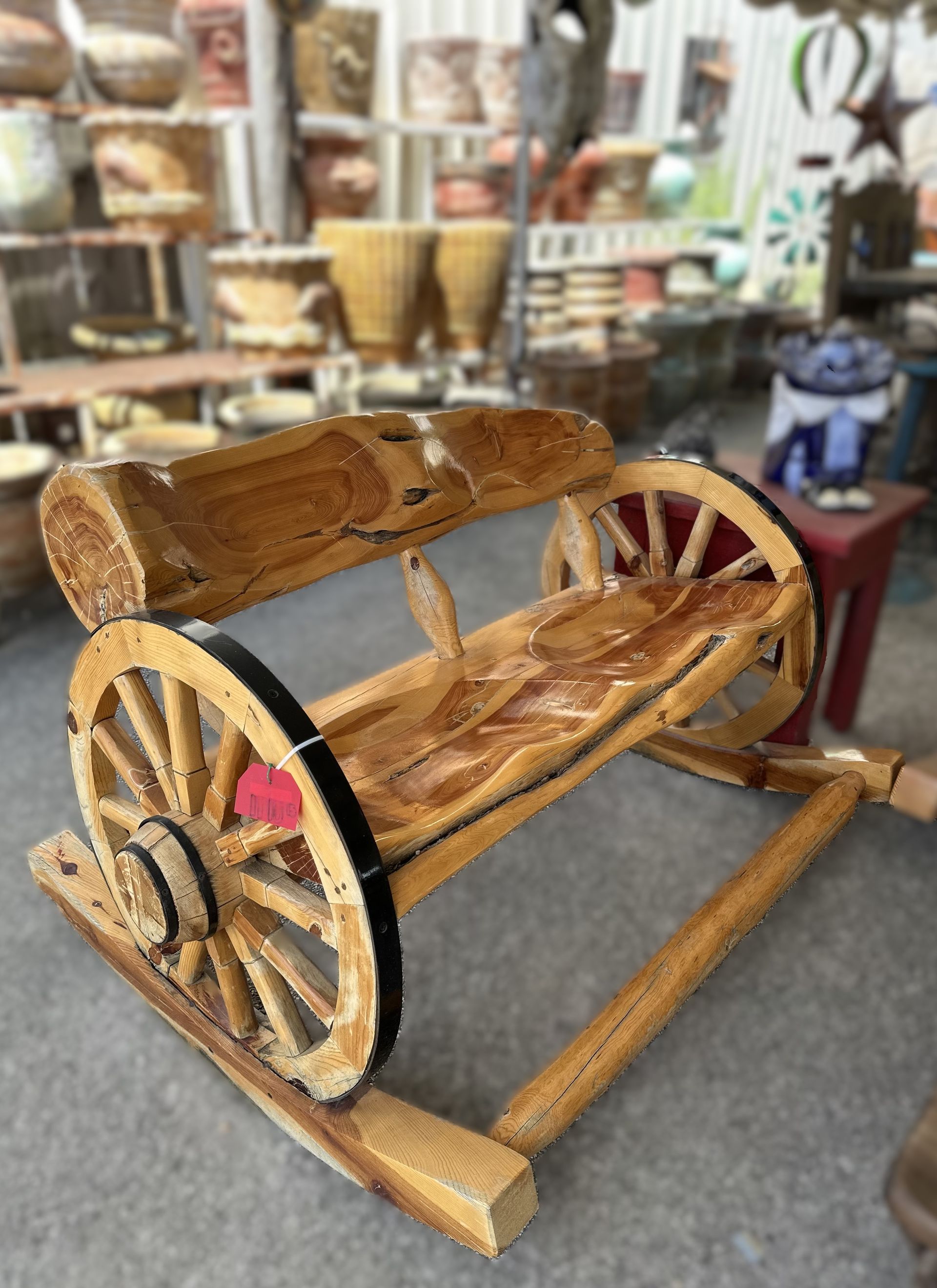 Wooden wagon wheel bench with a log back, displayed in a shop with pottery and other items in the background.