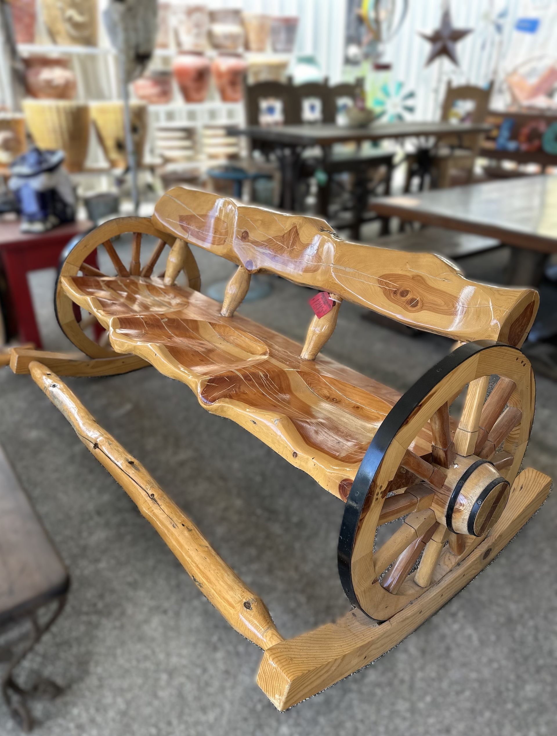 Wooden wagon-shaped bench with large spoked wheels, brown wood, and a red decorative element. It's inside a shop, with more furniture visible.