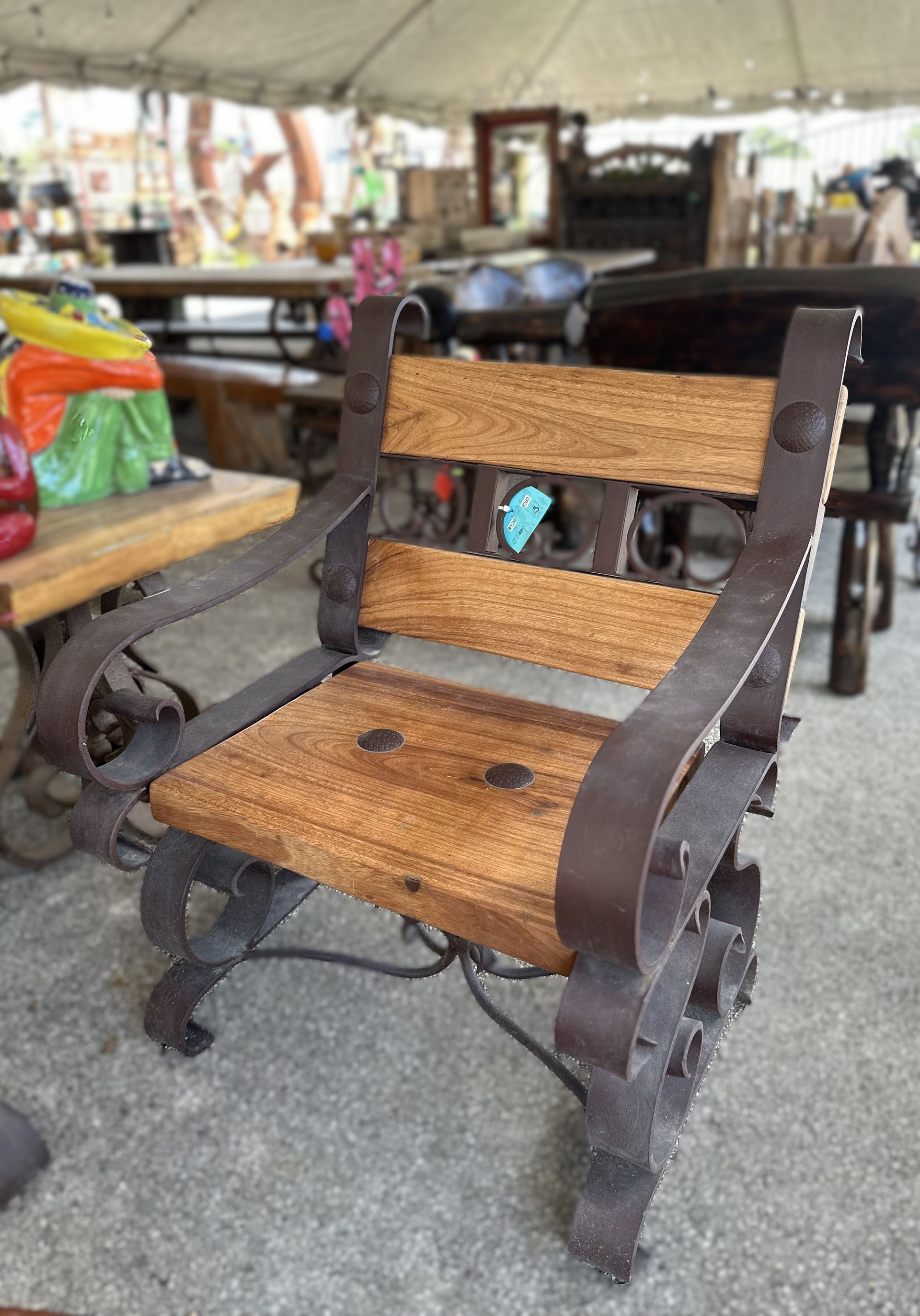 A rustic wooden chair with dark metal framing, set outdoors, likely for sale. The chair has a natural wood seat and back.