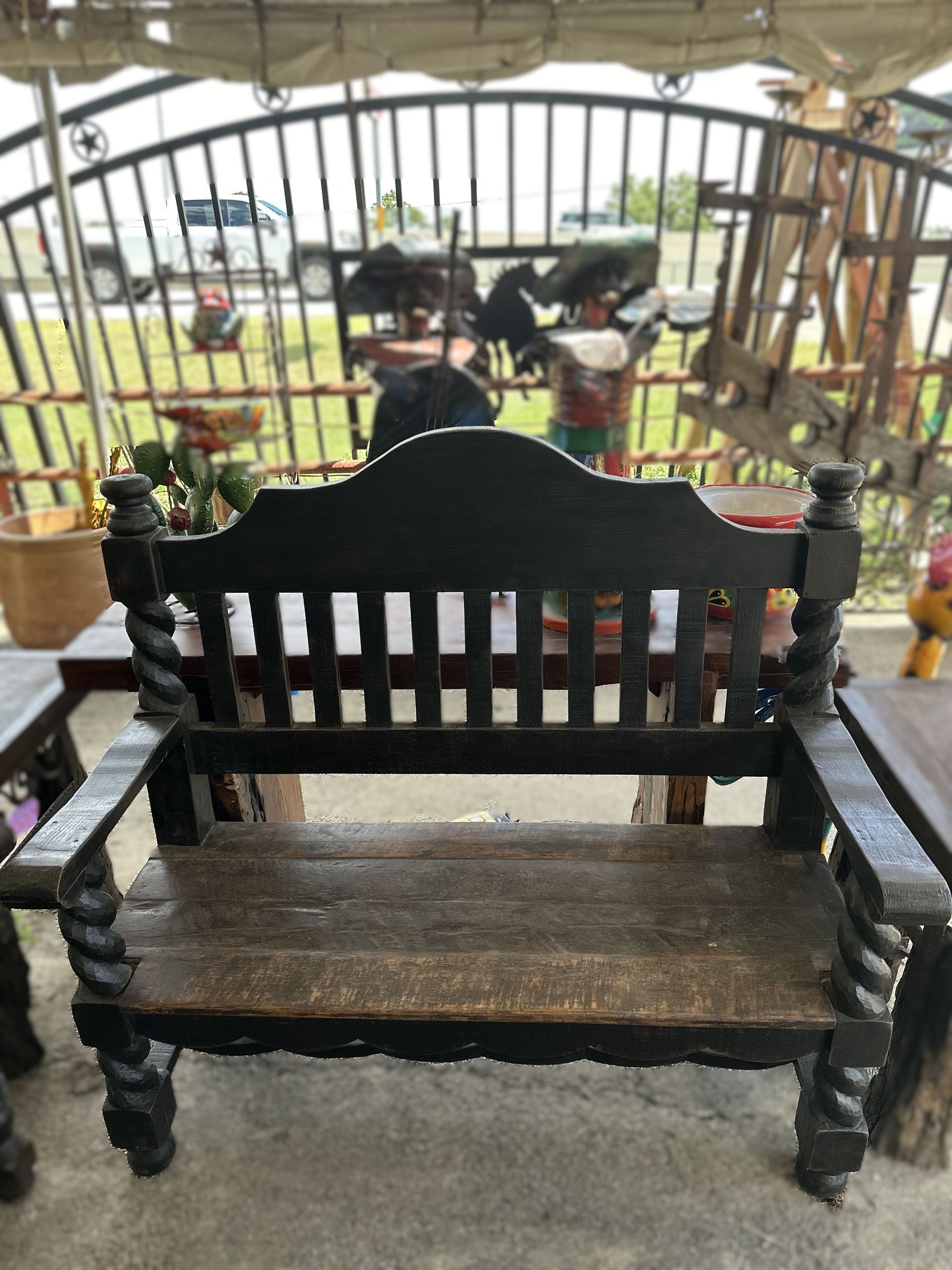 Black wooden bench with scrolled armrests and back, set outdoors near a fence and other items.