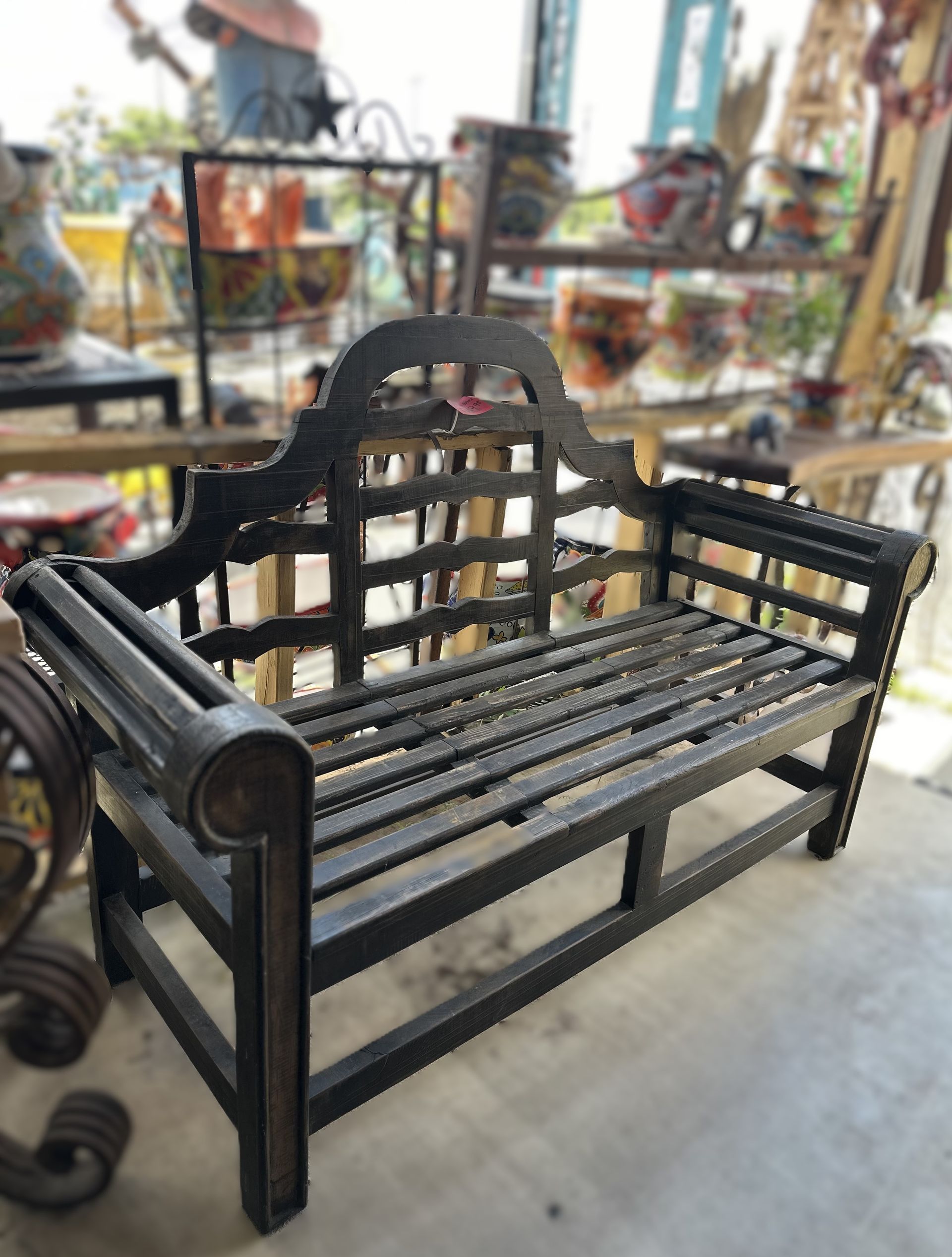 Black metal outdoor bench with slat seat and decorative back, on display in a shop with pottery in background.