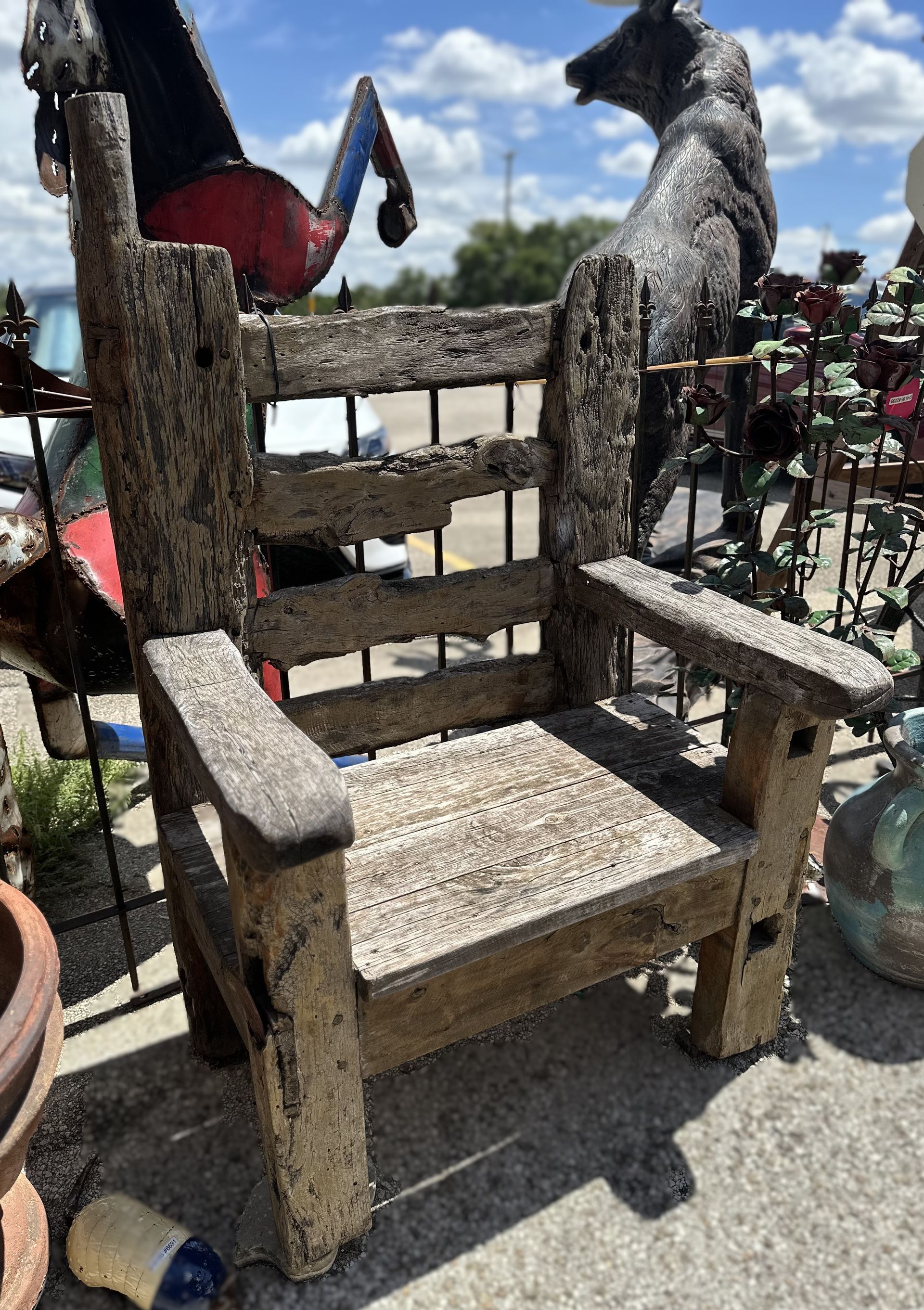 A weathered wooden chair with armrests and back slats sits outside. Other yard art items are visible in the background.