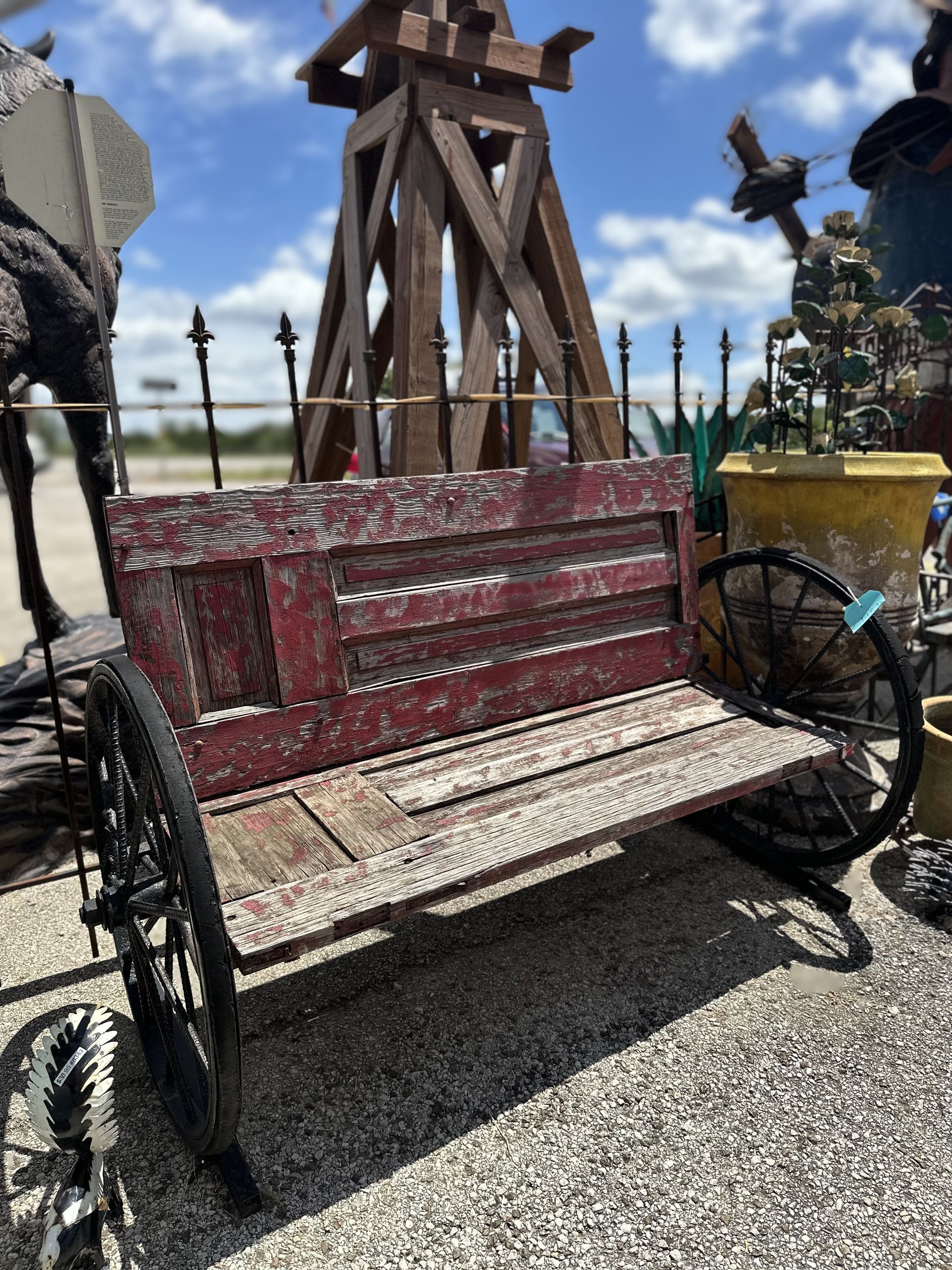 A rustic bench made from repurposed wood and metal, possibly from a wagon, sits outdoors. The bench is reddish-brown, and the background is blurry.
