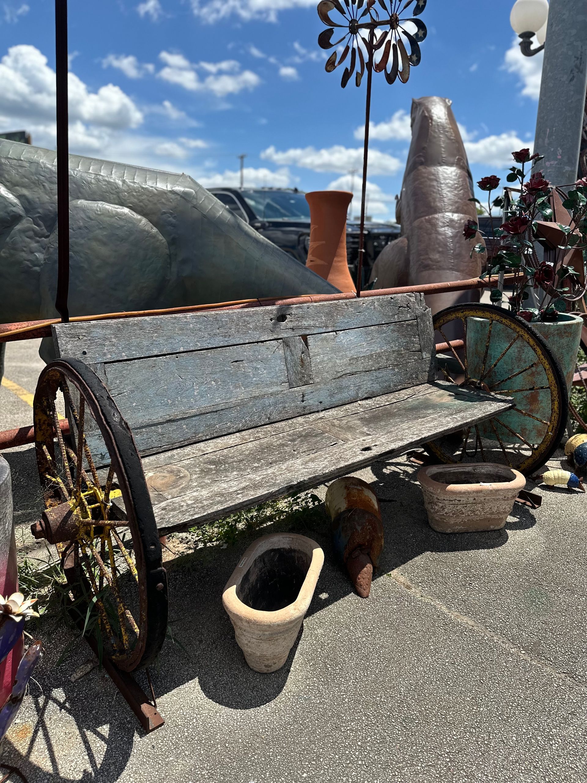 Rustic wooden bench with wagon wheel sides, surrounded by pottery and garden art. Blue sky background.