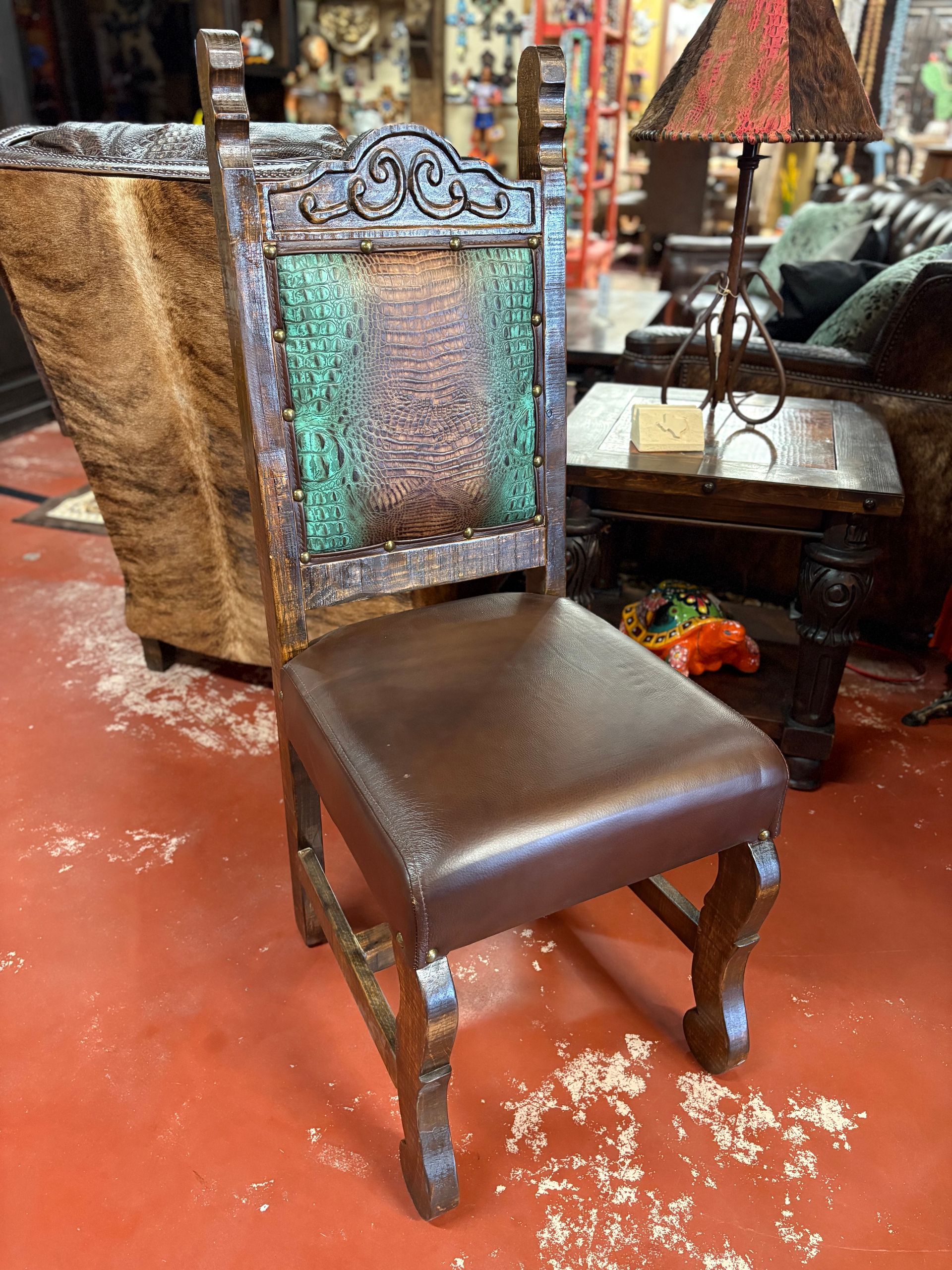 Carved wooden chair with a brown leather seat and a textured, colorful backrest. The chair sits on a red floor in a shop.