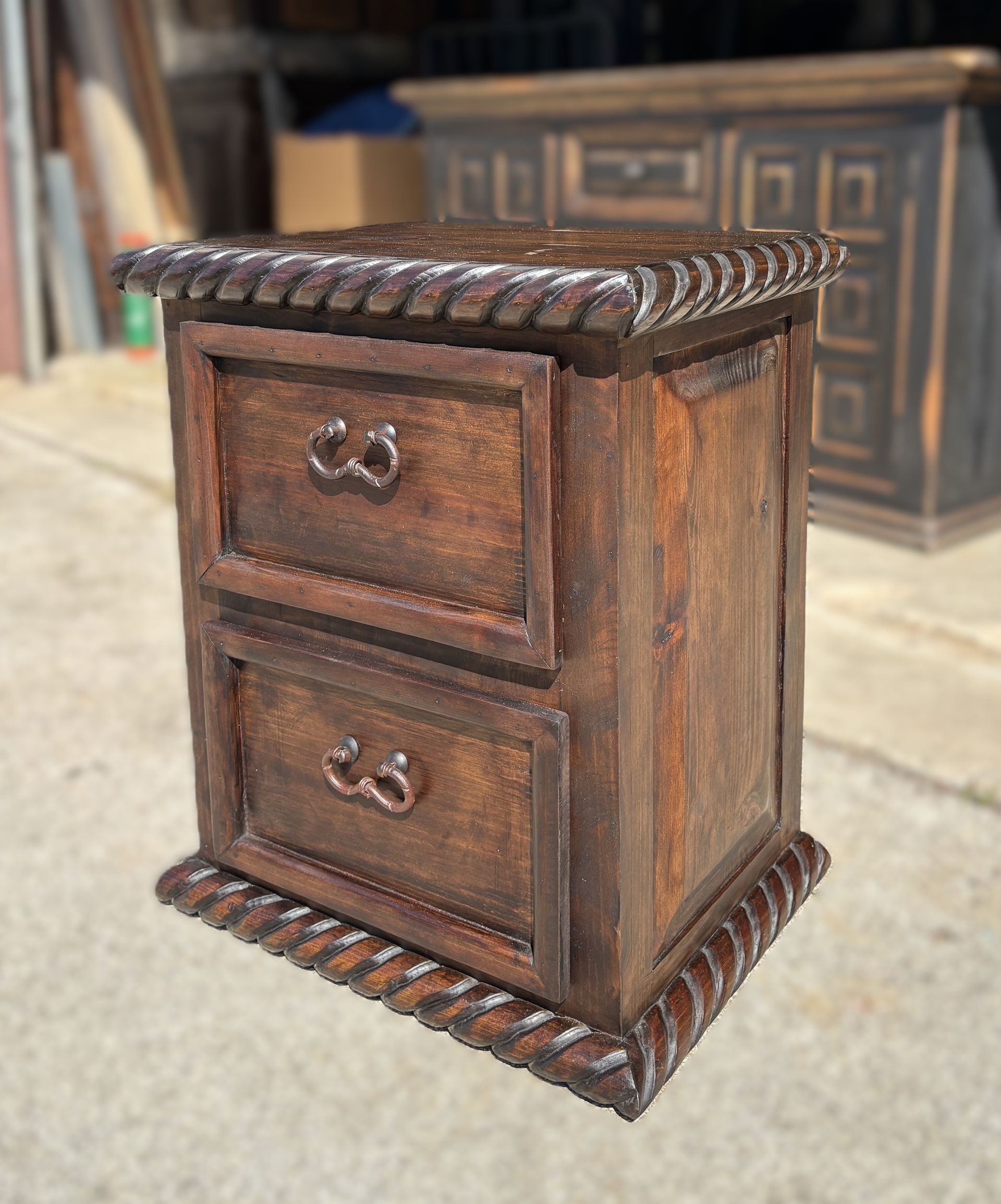 Wooden nightstand with two drawers, dark brown, rope trim on top and bottom, outdoors.