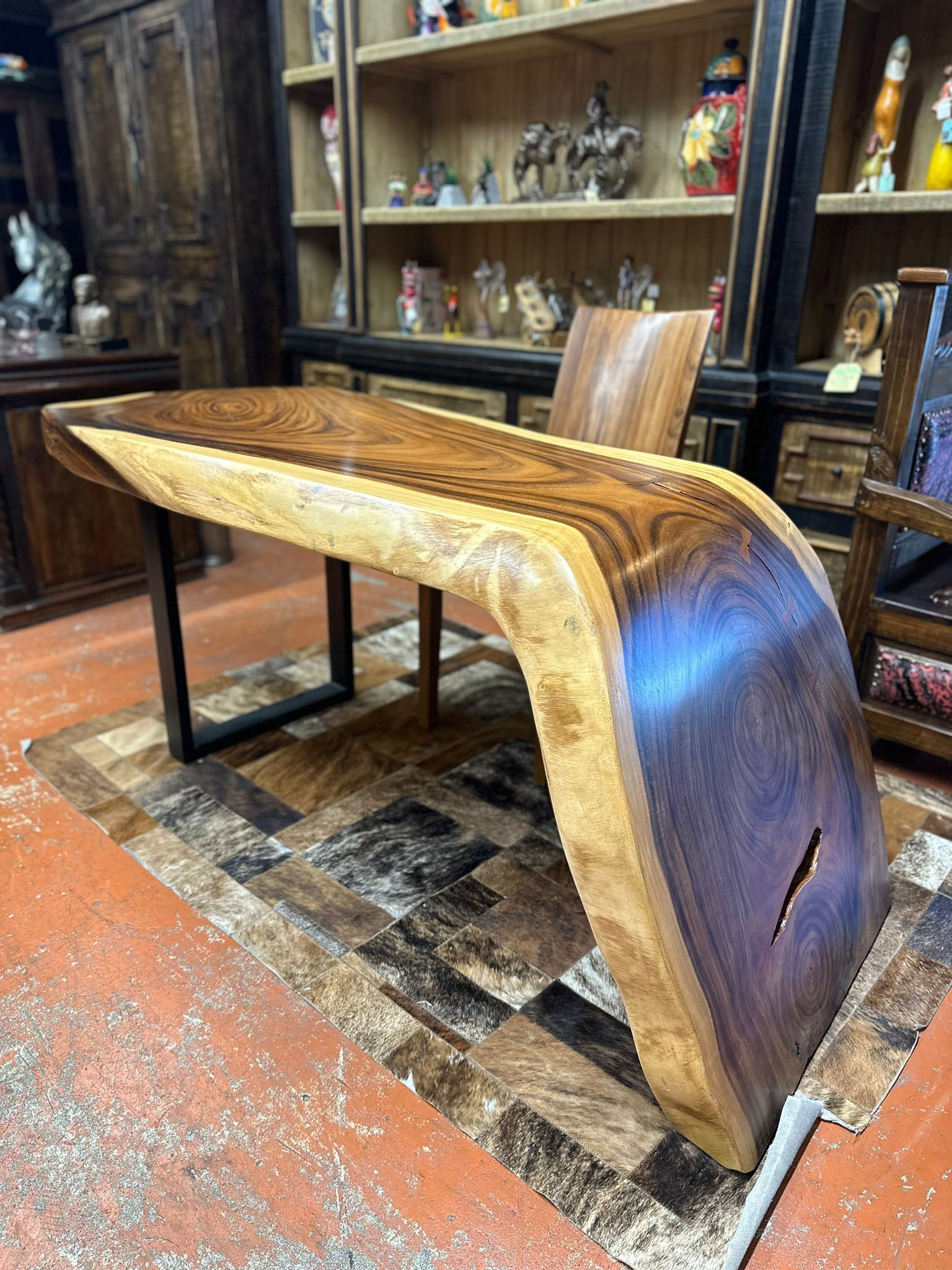 Live-edge wooden desk with dark wood finish and metal legs on a patterned rug, with shelves and chair in the background.
