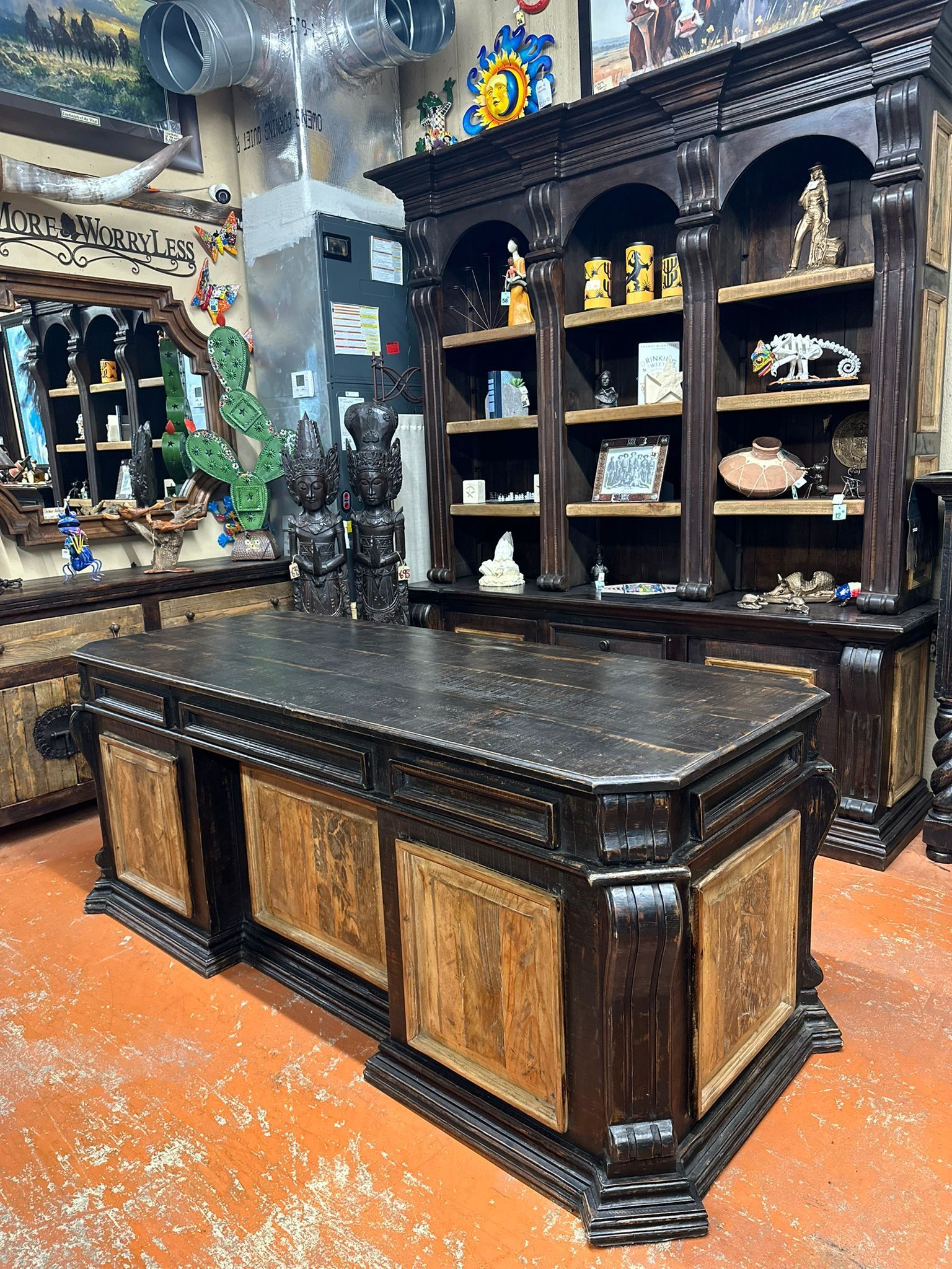 A dark wooden desk and matching bookshelf are in a room. The desk has ornate carvings and a textured surface. The shelves are filled with decorative items.