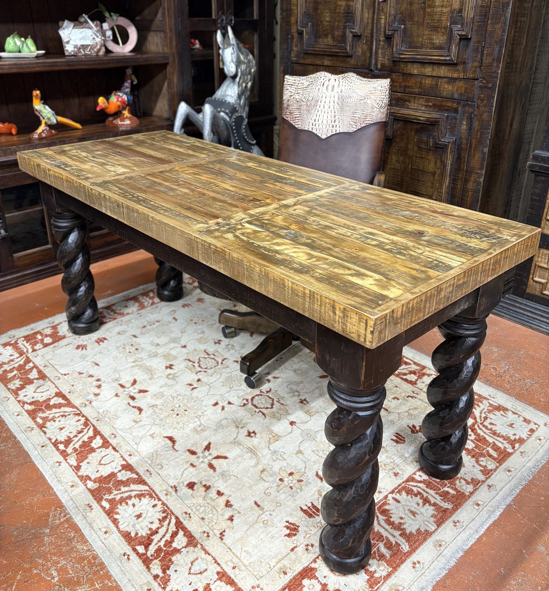 A miniature wooden desk with twisted legs sits on a patterned rug in a room with a bookshelf.