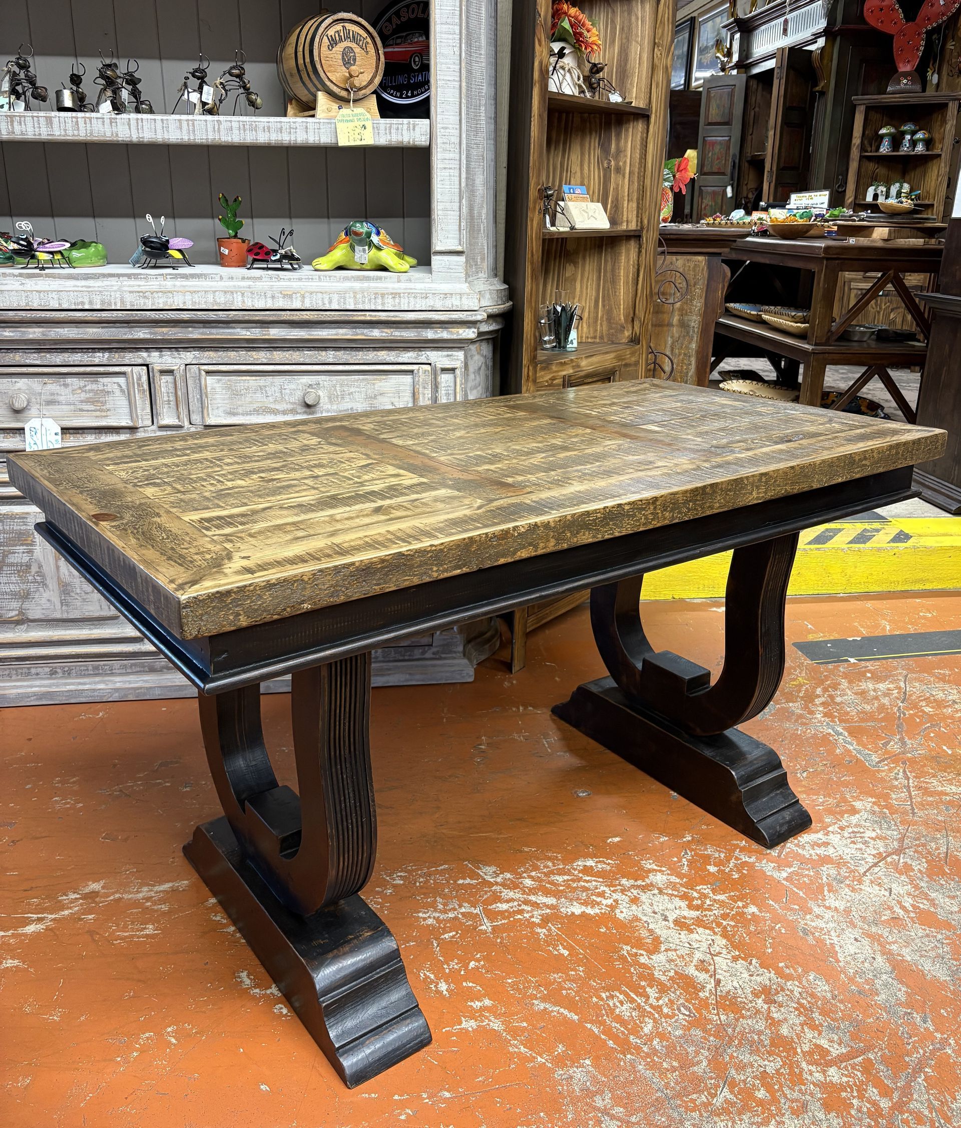 A rustic wooden desk with a light brown top and black decorative legs, inside a store on an orange floor.