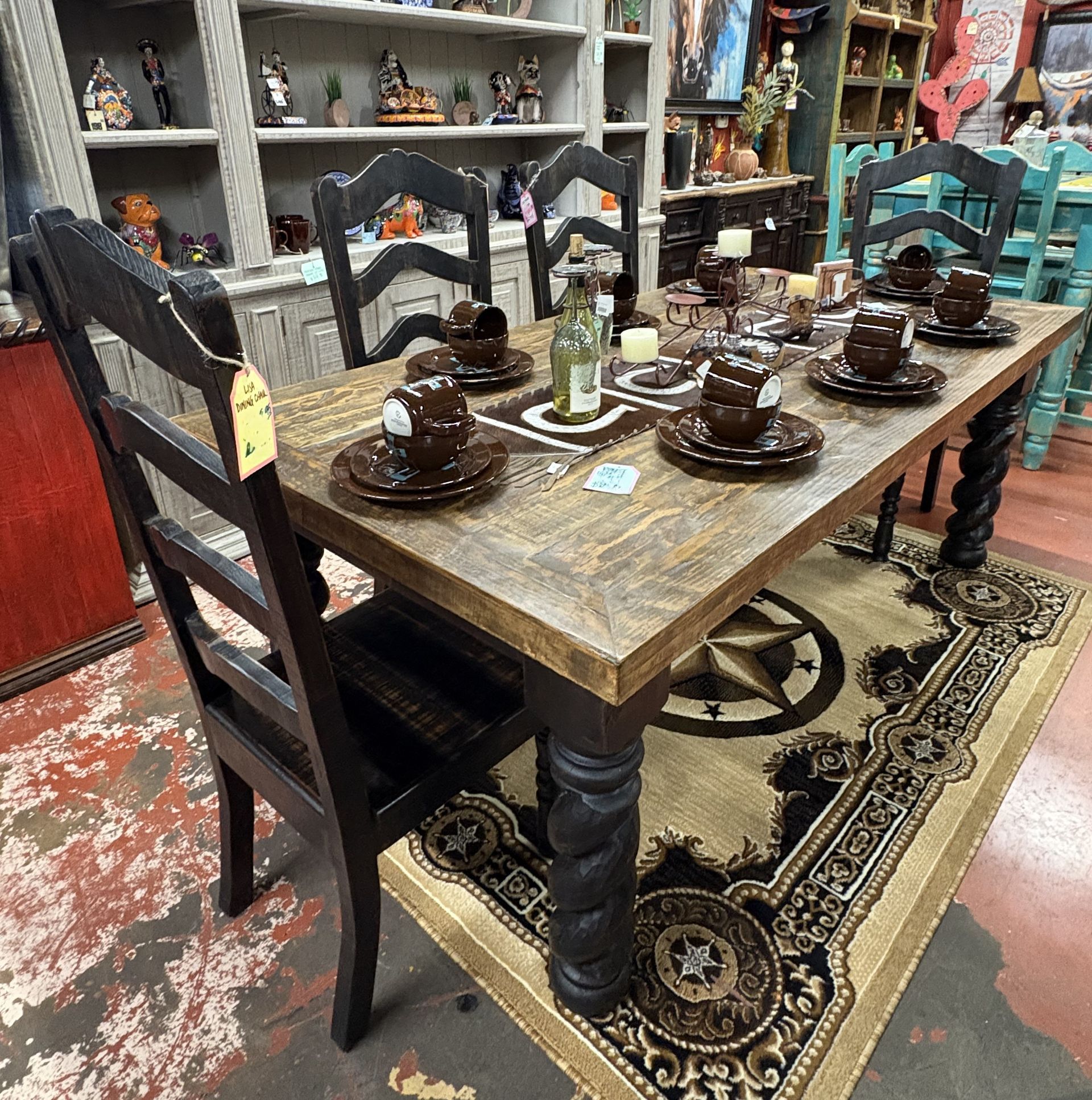 A wooden dining table with a rustic rug, set for a meal. Black chairs surround the table, in a shop setting.