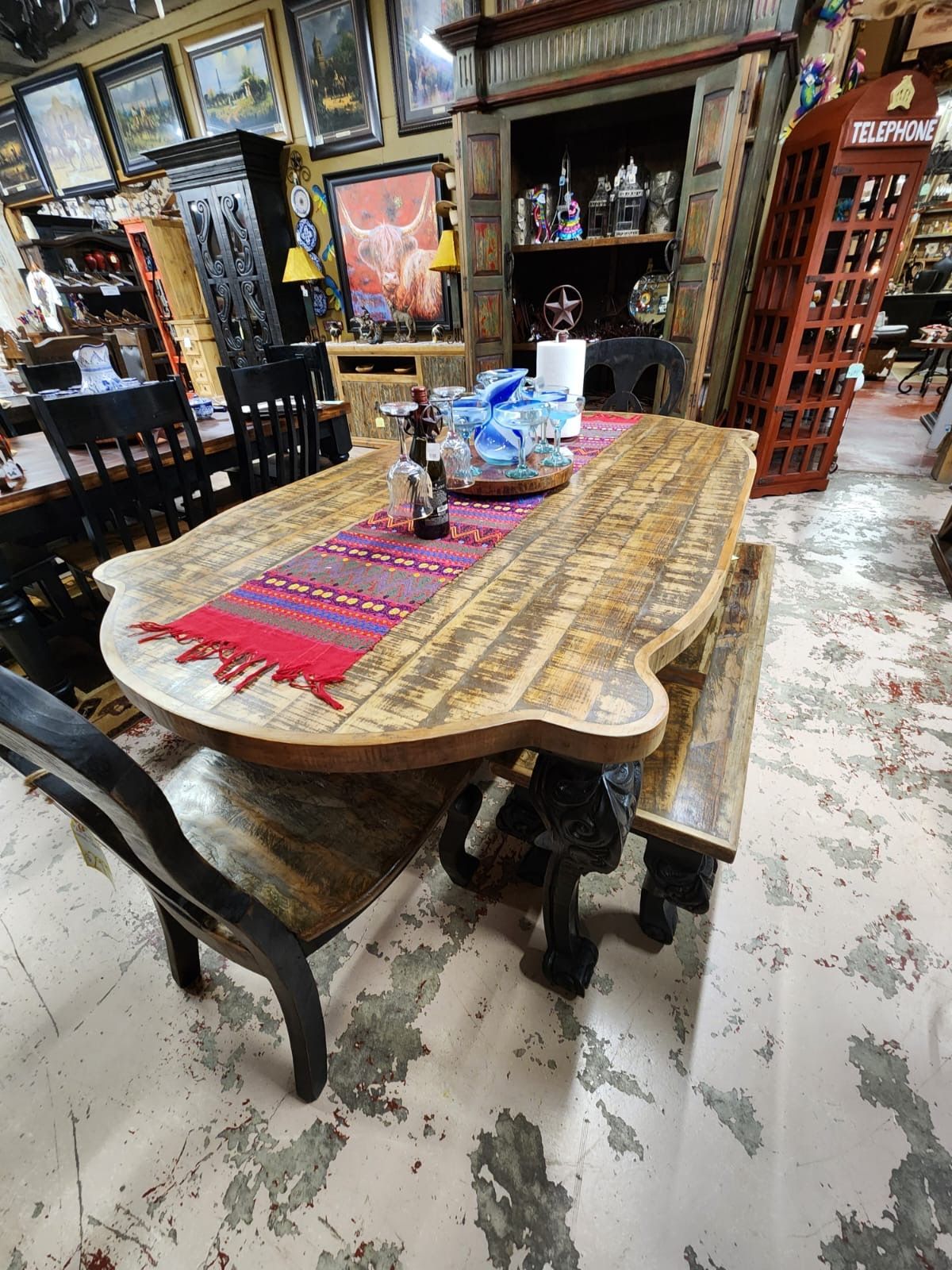 Rustic wooden dining table with matching bench and chair, set in a cluttered antique shop. Table has a colorful runner.