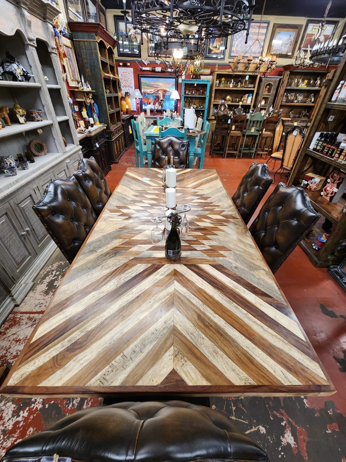 Long, wooden dining table with a herringbone pattern and tufted leather chairs in a well-stocked store. Candles sit in the center.