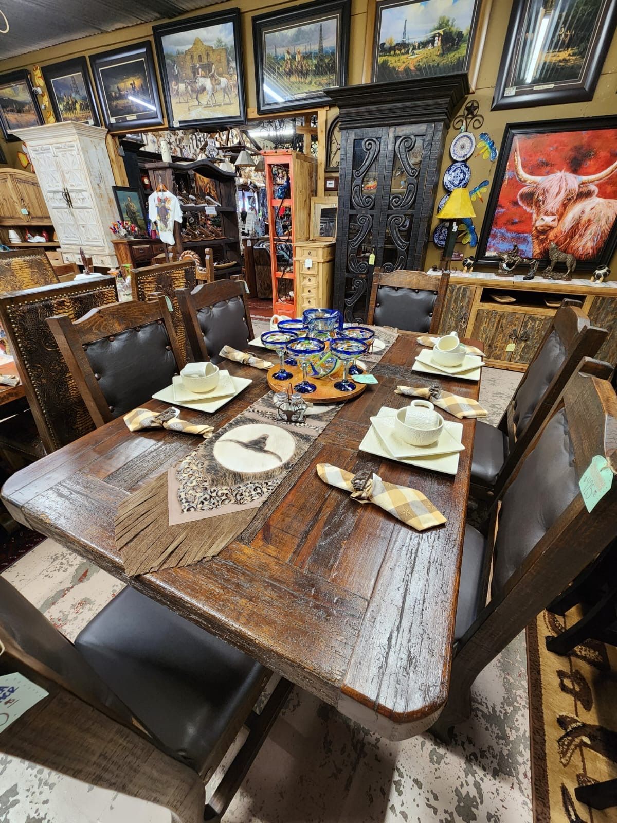 Dining table set in a rustic furniture store. The wooden table is set with plates and napkins, surrounded by leather-seated chairs.
