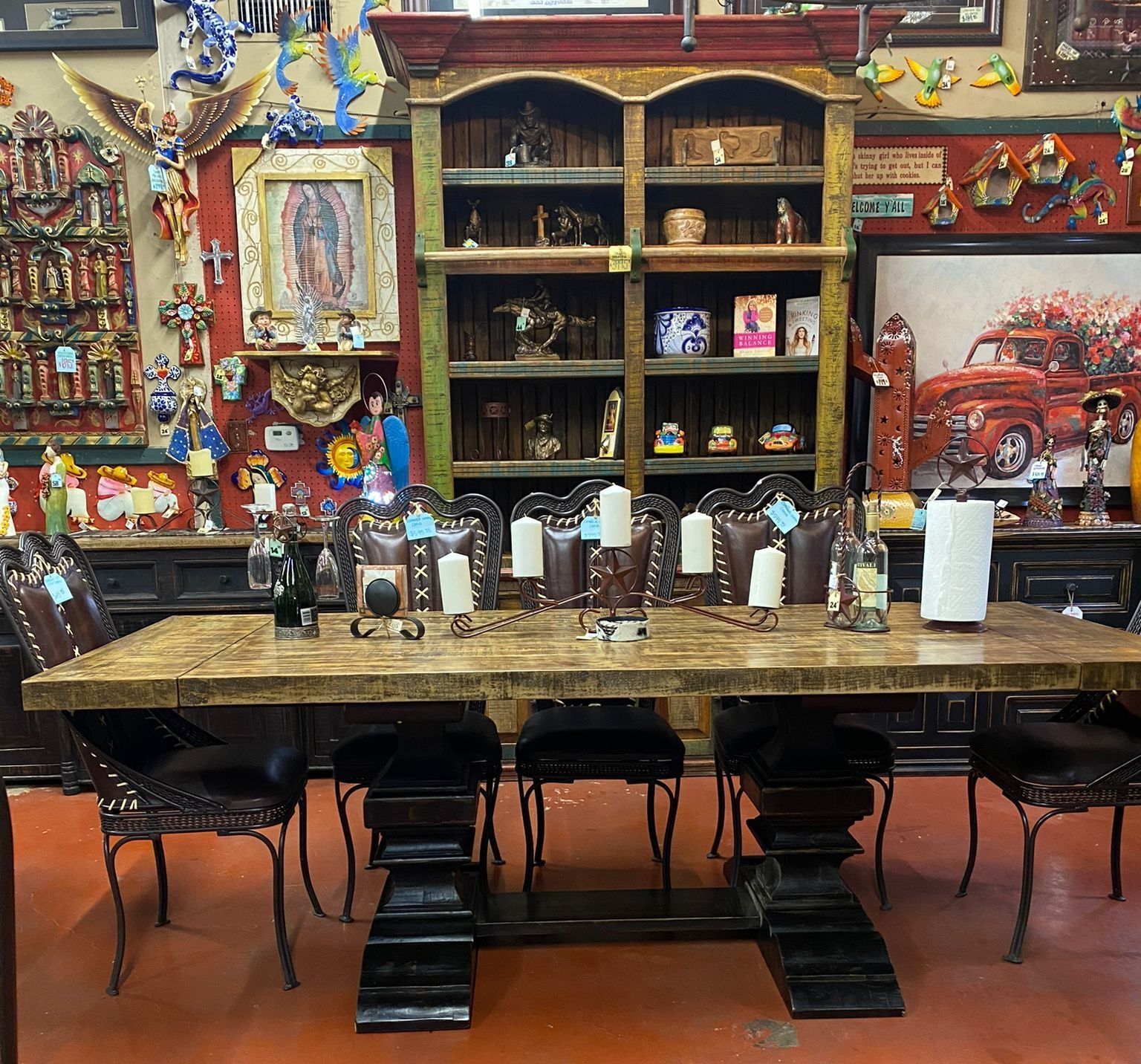 A dining room set with a large wooden table, leather chairs, and candle holders, set against a colorful wall with art and shelves.