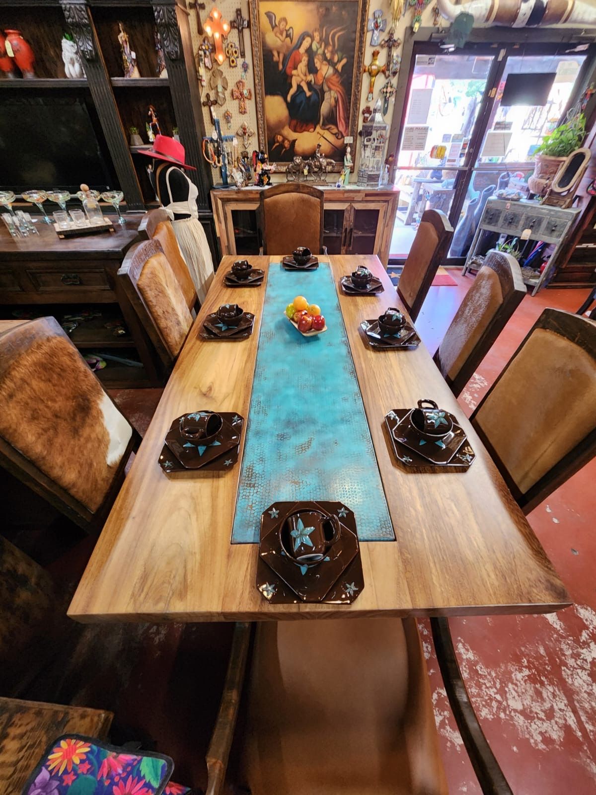 Long wooden dining table with place settings, teal runner, and surrounding chairs in a store.