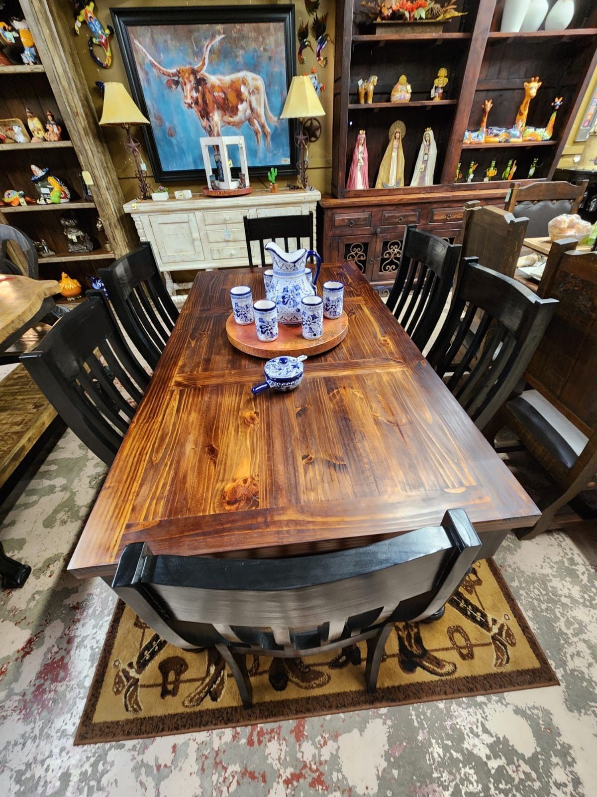 A wooden dining table with six black chairs, centered on a rug with cowboy boot design. A painting of a longhorn is on the wall.