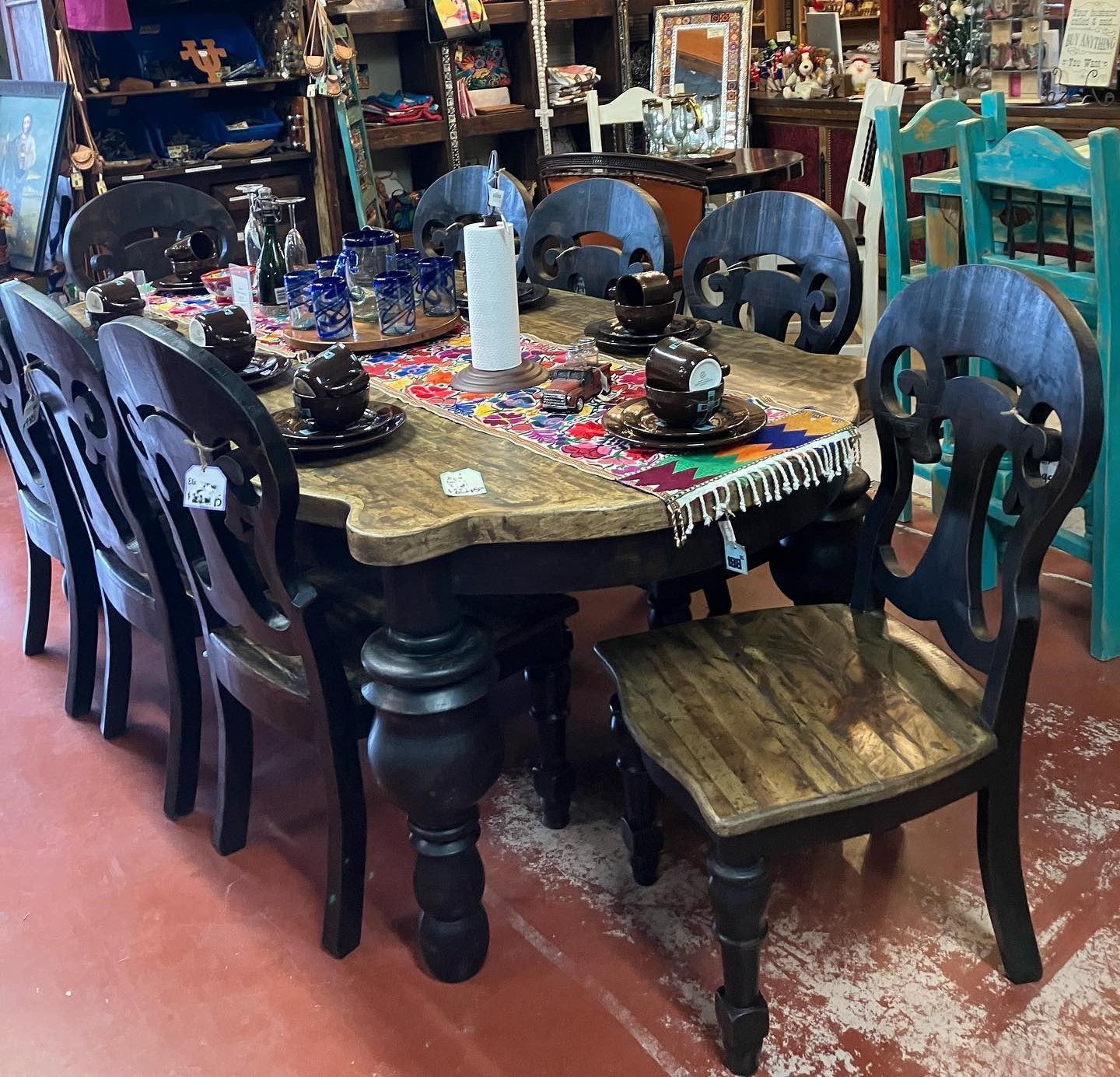 Dining table with six chairs, dark wood with ornate backs. Table set with place settings and colorful tablecloth, inside a store.