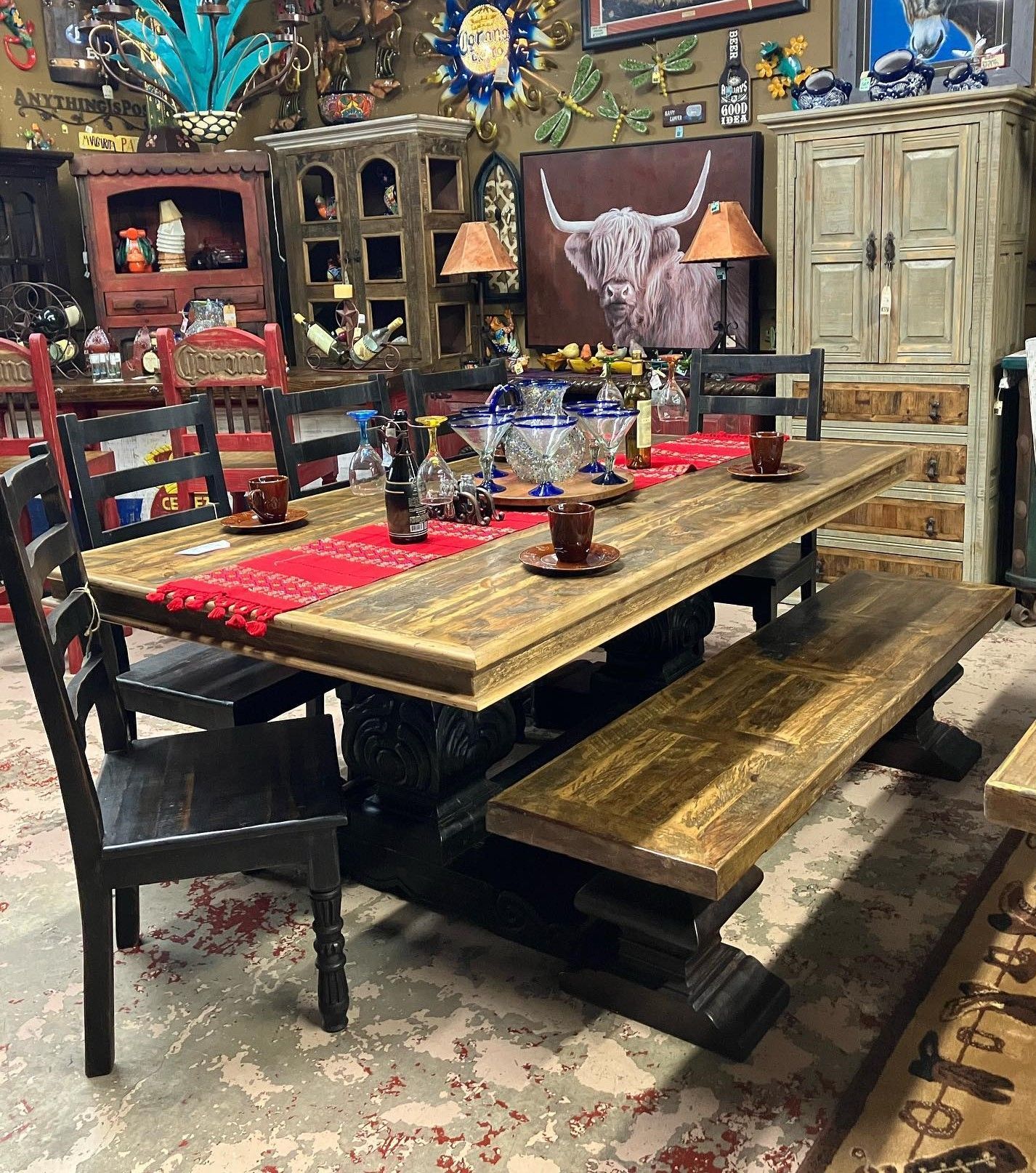 A rustic wooden dining table with chairs and a bench in a cluttered interior setting.