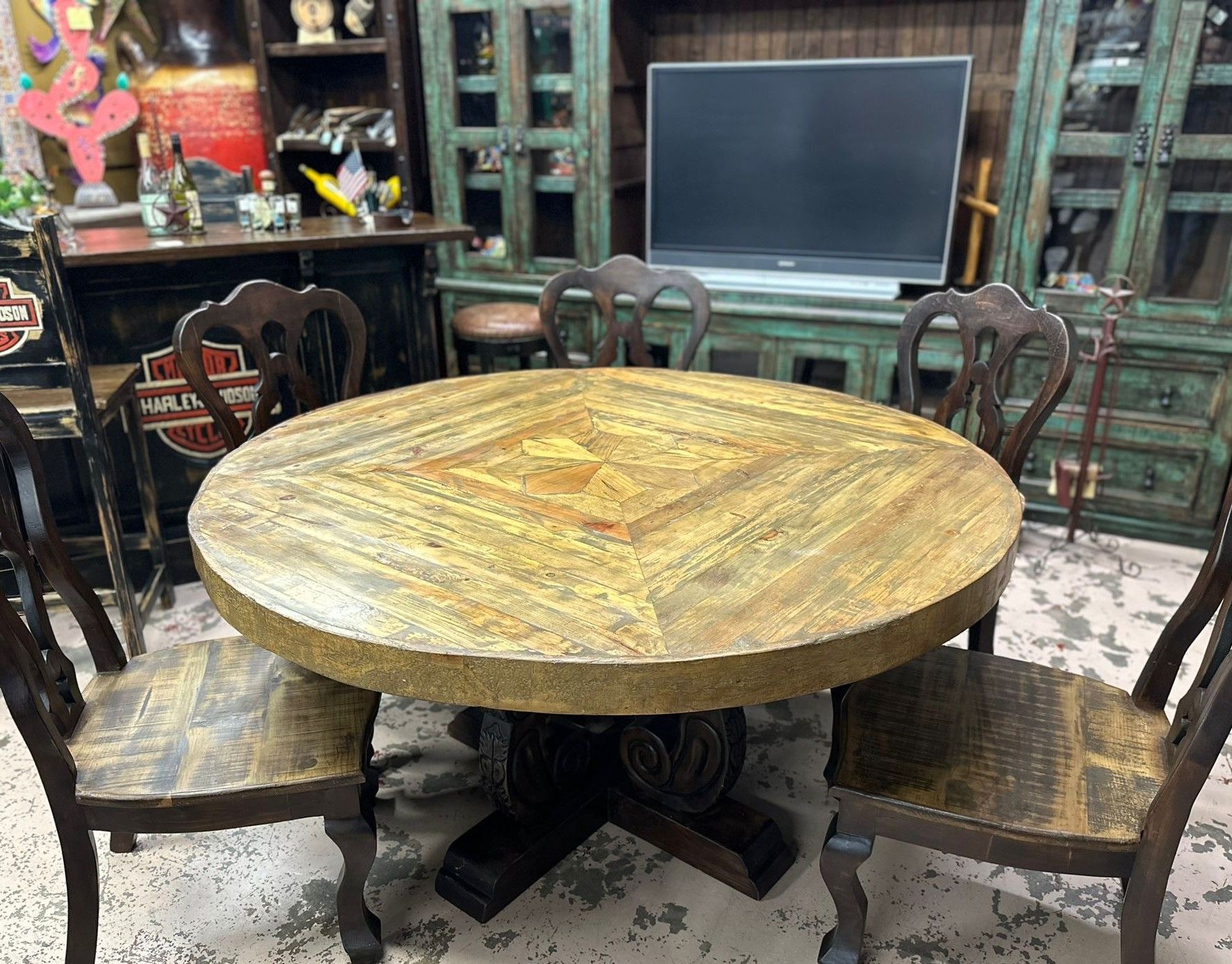 A round, rustic wooden dining table and chairs set in a room with decorative furniture. The table is light brown and the chairs are dark brown.