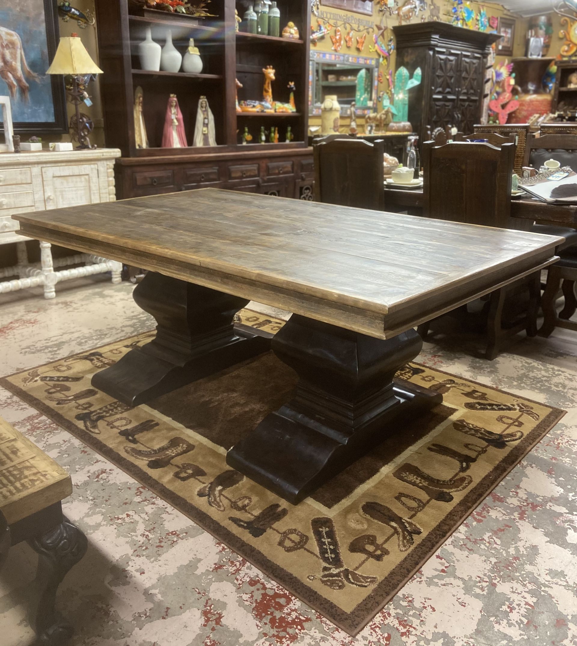 A large wooden dining table with dark pedestal bases sits on a patterned rug. Shelves and other furniture are in the background.