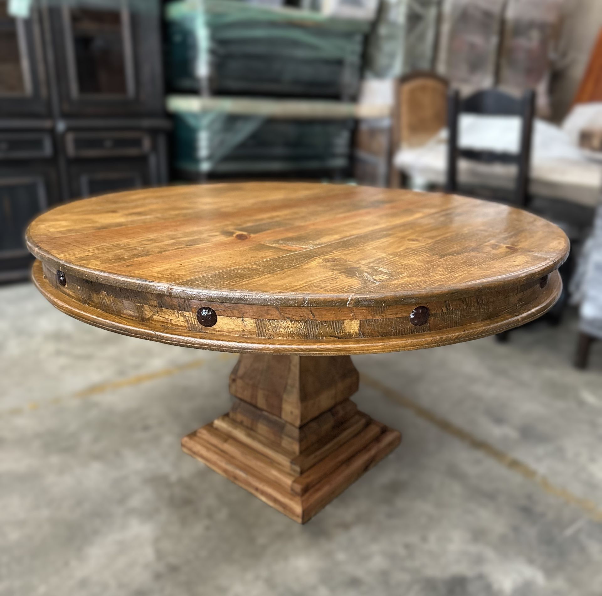 Round wooden dining table with a pedestal base, brown tones, and studded rim, in a furniture store.