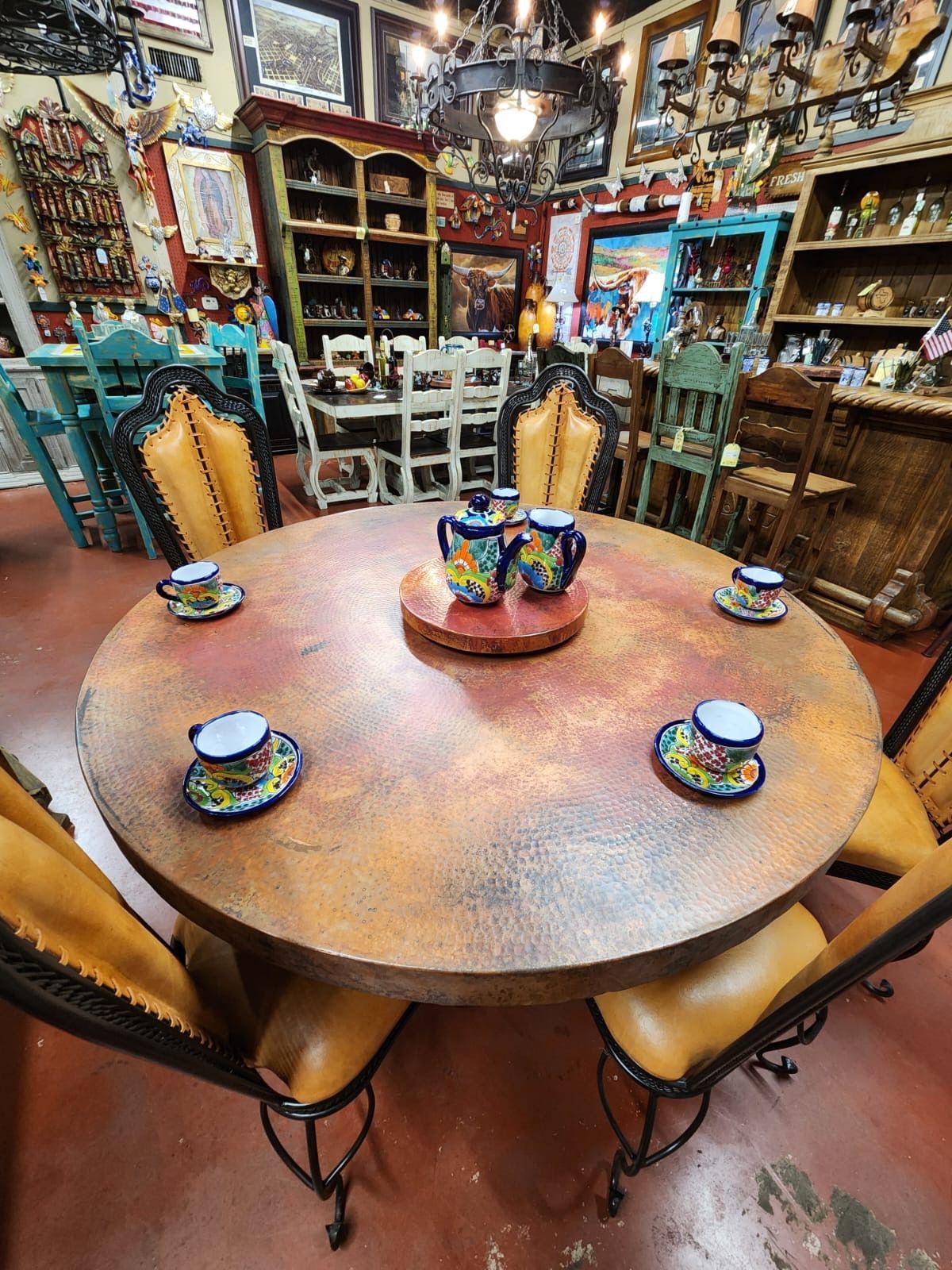 Round copper-toned table with colorful teacups, surrounded by tan leather chairs. Inside a shop filled with rustic decor.
