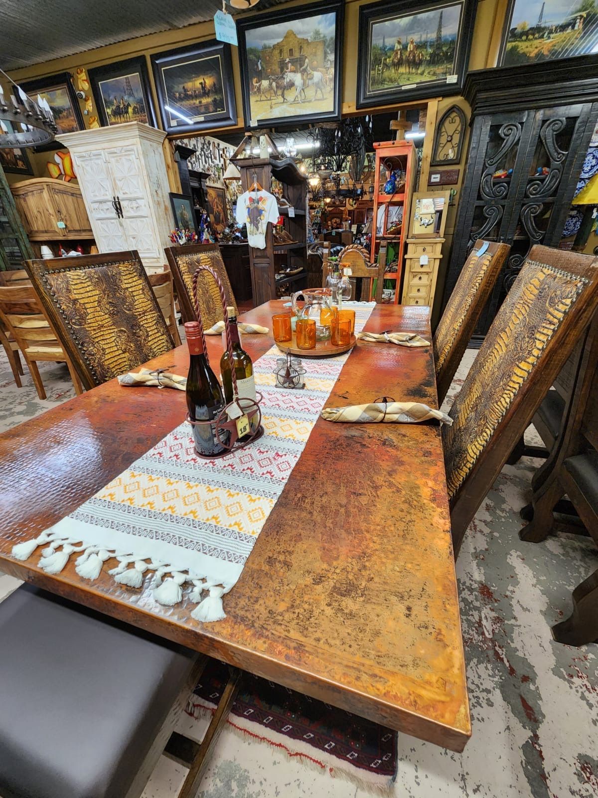 A copper-topped dining table with six patterned chairs, set with a runner, bottles, and drinks in a shop.