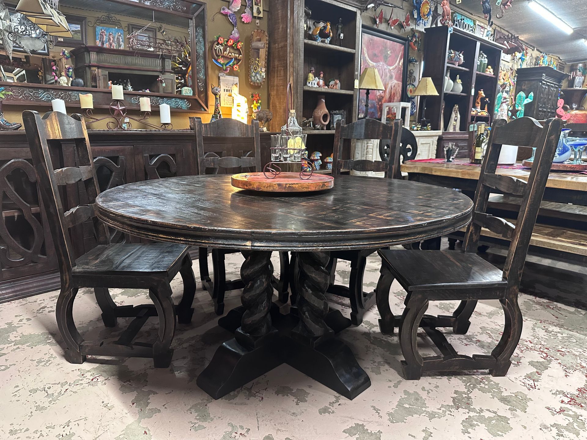 A dark wooden round dining table with spiral base and matching chairs. The setting is a shop filled with other rustic furniture.