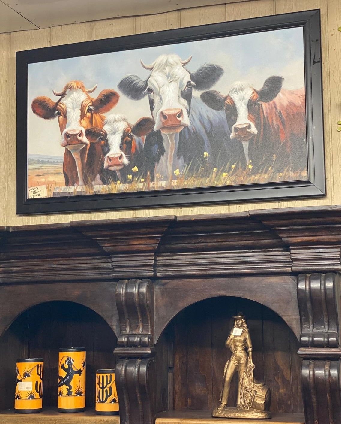 Painting of five cows looking forward in a field, framed above a dark wooden shelf with decor.