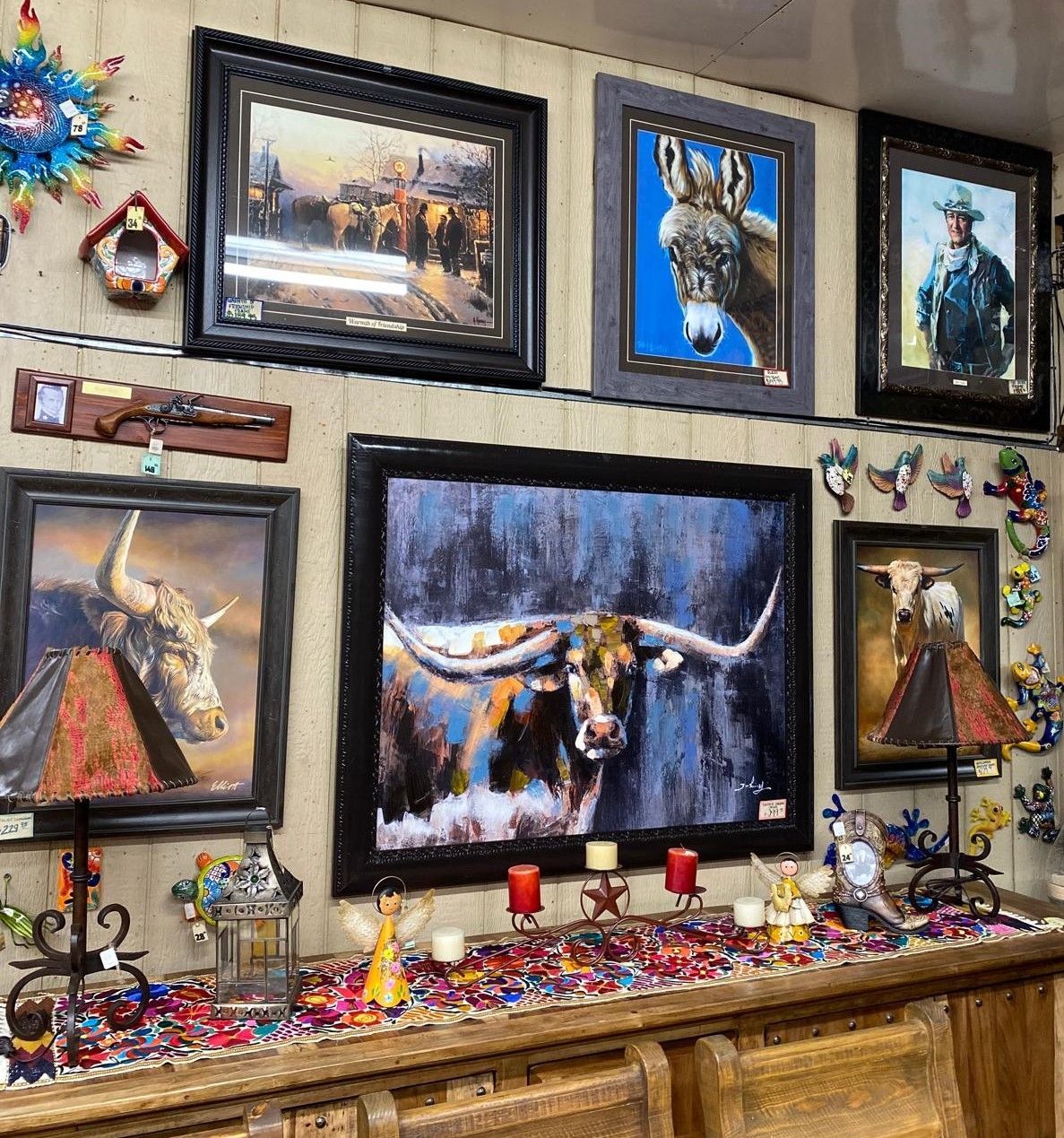 Display of framed art featuring western themes, including a longhorn, donkey, and cowboys.  Wooden furniture with colorful decorations is in the foreground.