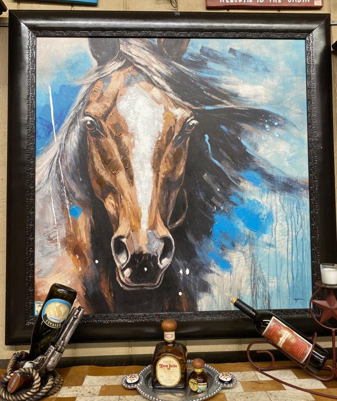 A painting of a horse with a white blaze and brown muzzle is framed with decor including tequila and wine bottles.