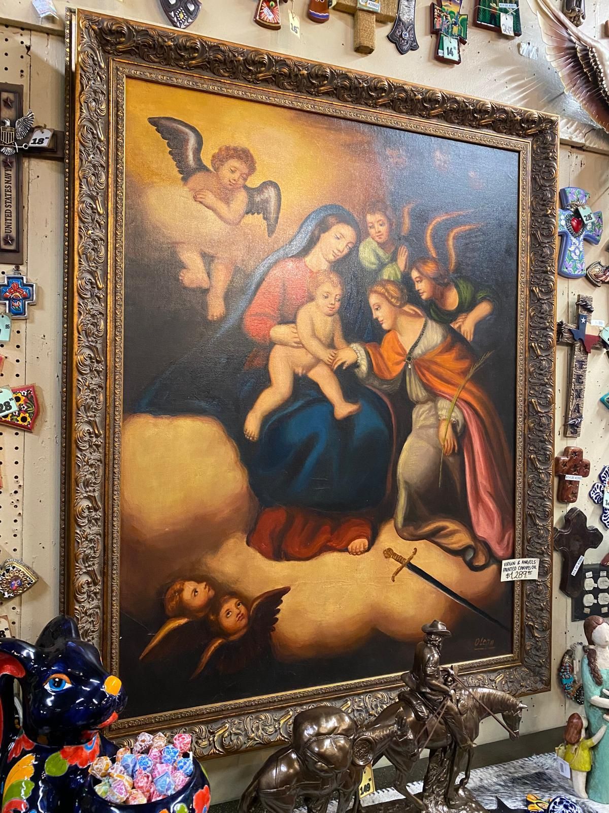 Painting of Mary holding baby Jesus, surrounded by angels, framed in gold. Scene set in clouds, other religious objects visible nearby.