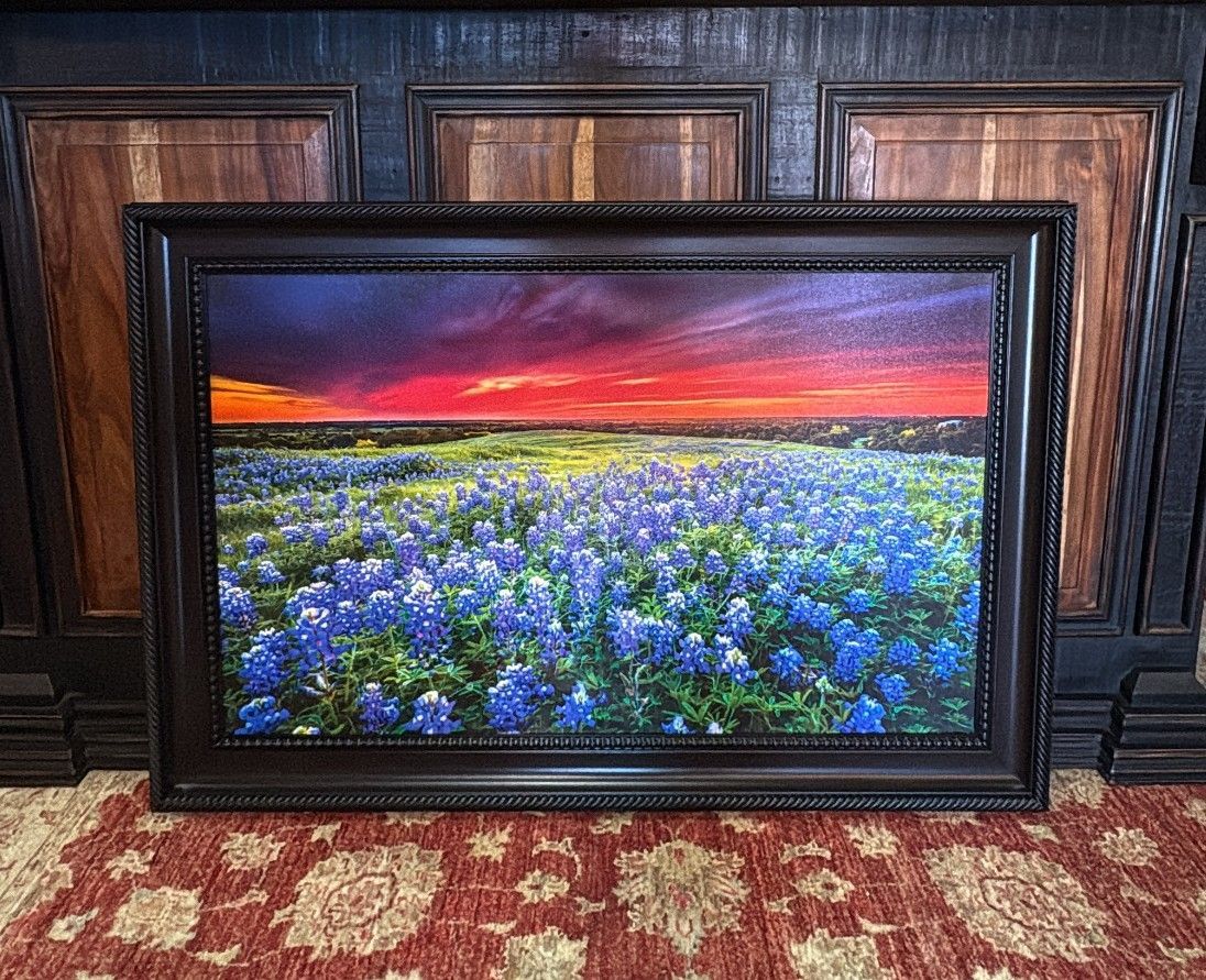 A framed painting of a Texas bluebonnet field at sunset with a vibrant orange and purple sky.