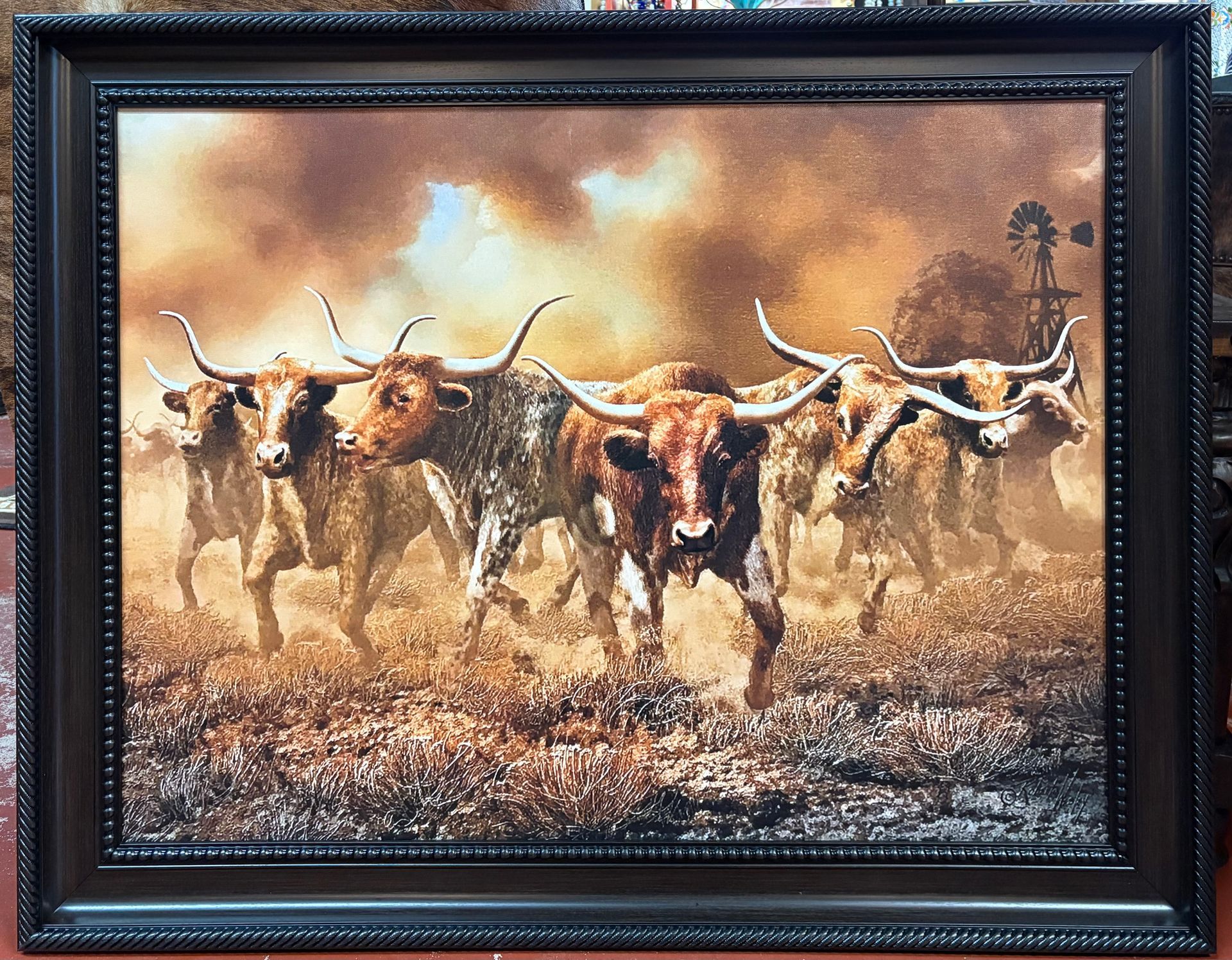 Longhorn cattle running through a dusty field under a cloudy sky, framed by a dark decorative border.