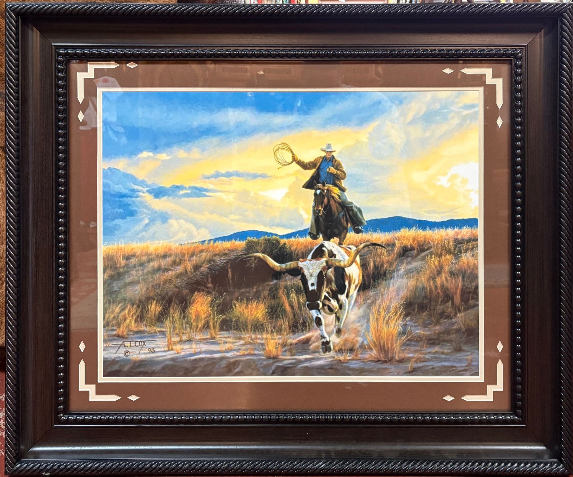 Cowboy on horseback roping a longhorn steer in a field under a blue sky with sunset hues. Painting in a dark wood frame.