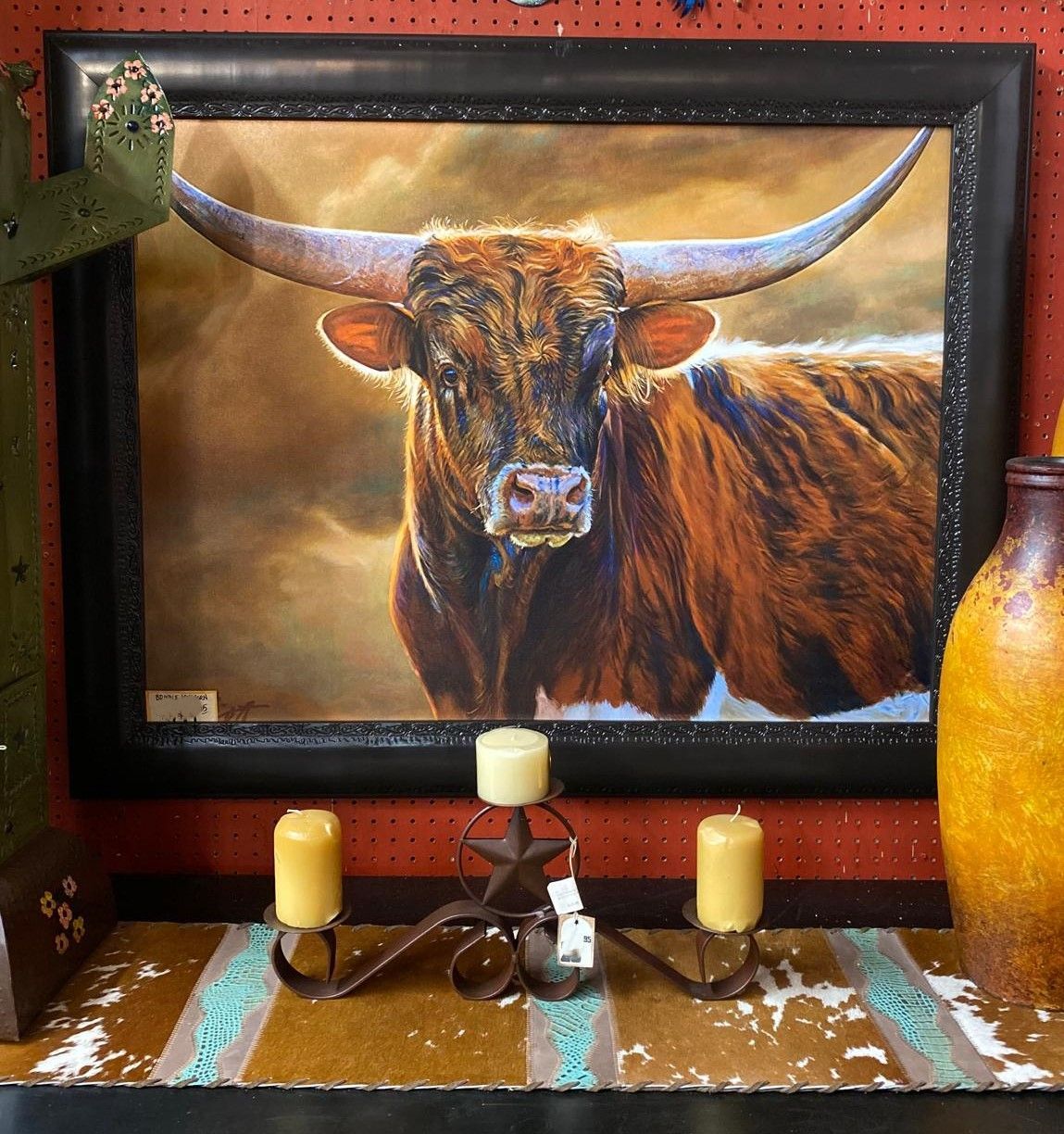 Framed painting of a longhorn bull with large horns. It rests on a shelf with candles and a decorative vase.